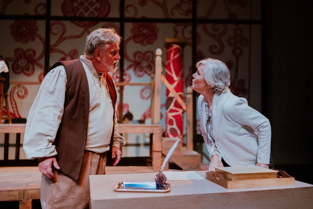 Two actors on stage in a dramatic scene, one wearing historical costume, standing near a desk with quill and paper, theatrical backdrop with floral patterns.