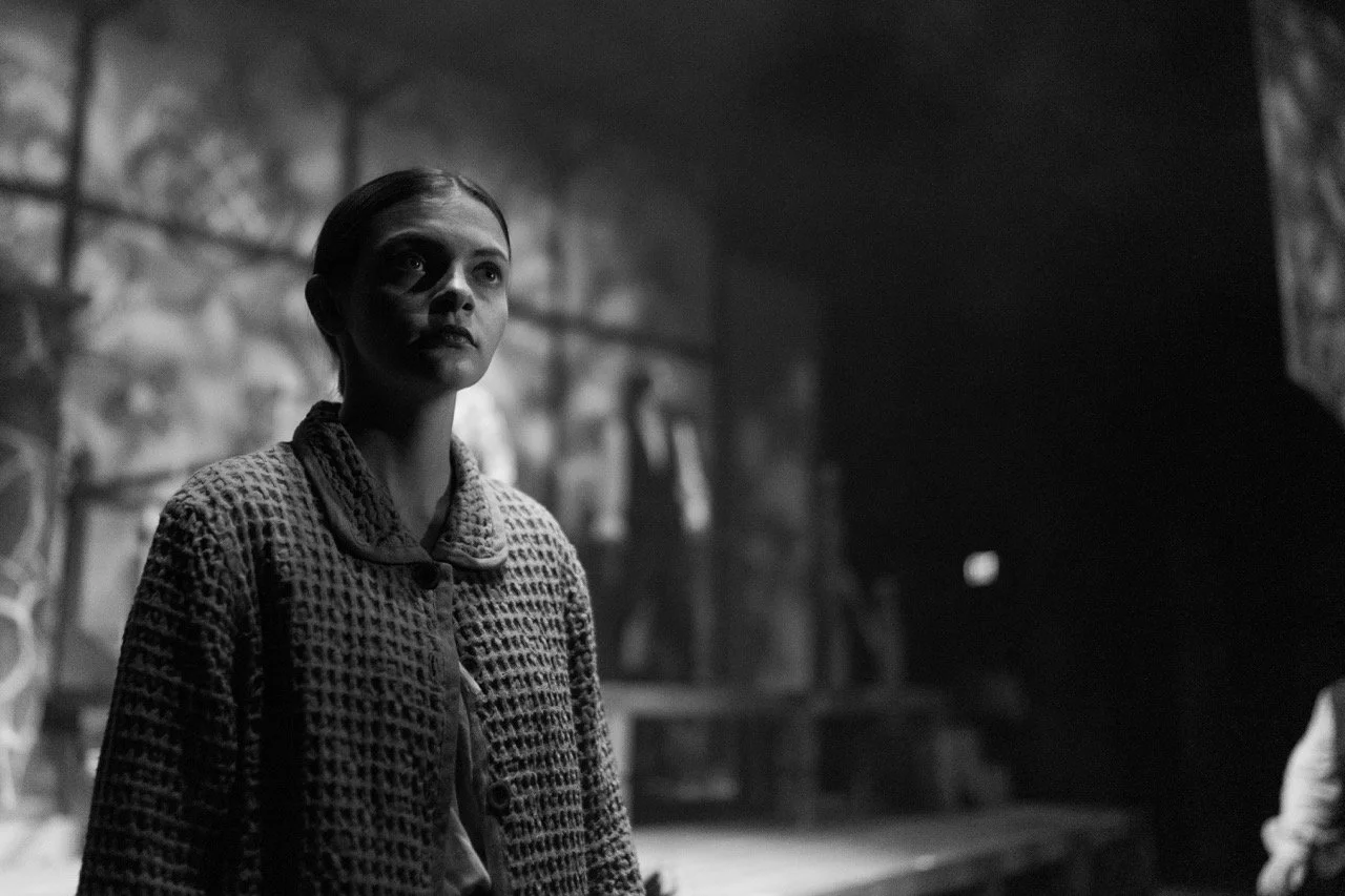 Black and white photo of a person in a patterned coat standing on a stage with dramatic lighting and blurred background.