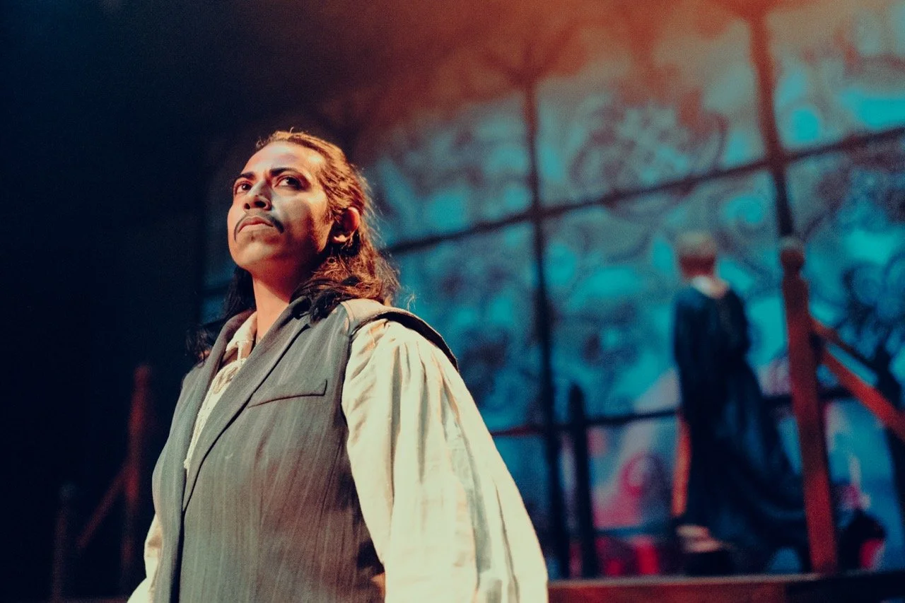 Person on stage in period costume with dramatic lighting, blurred background of ornate design.