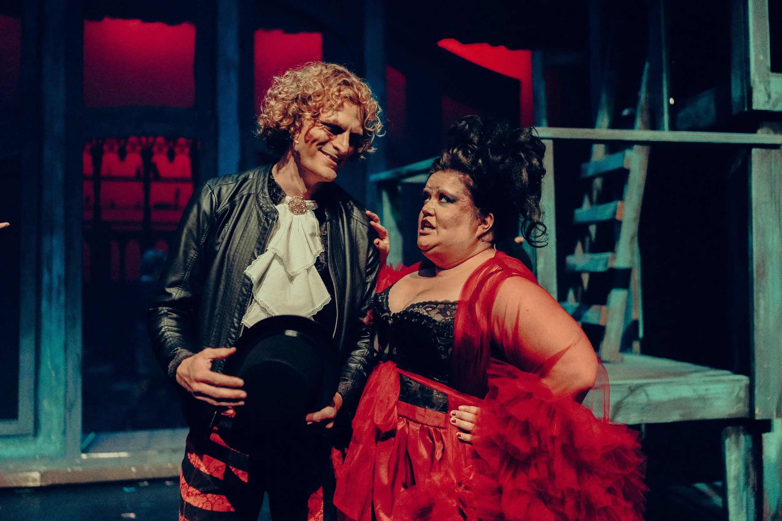 Two actors in theatrical costumes, one with curly hair holding a hat, and one wearing a red, ruffled dress, stand on stage with a dimly lit set in the background.