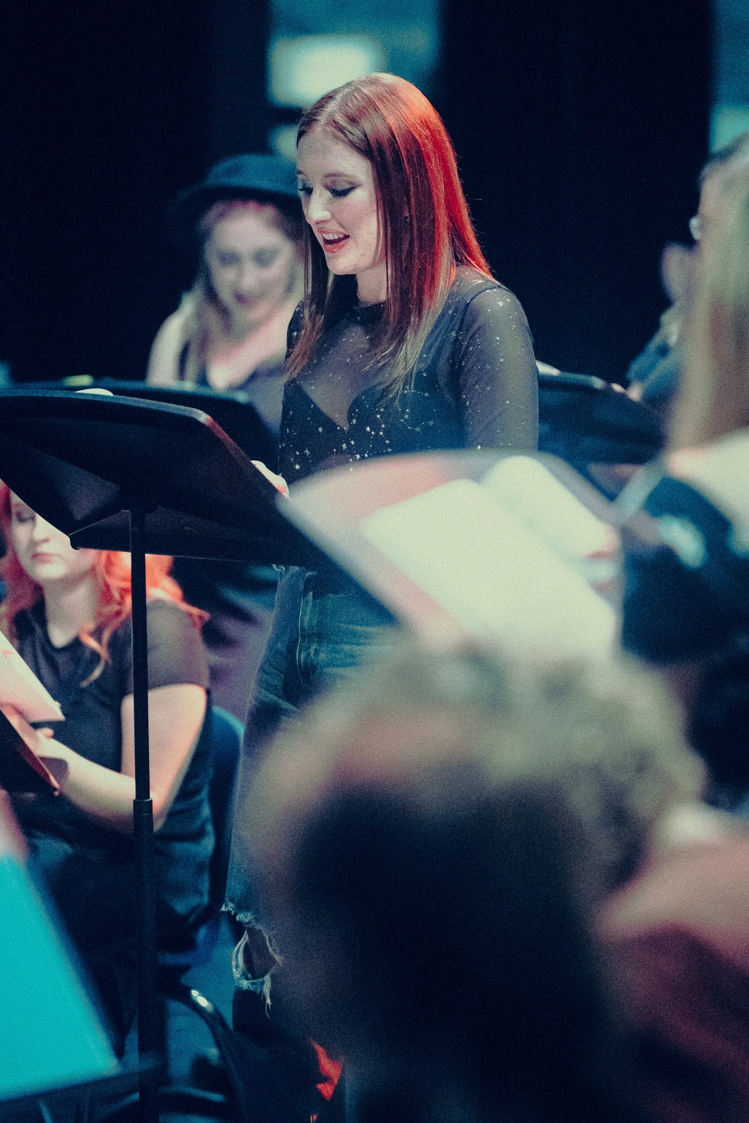 Woman with red hair reading from a music stand during a performance or rehearsal, with other people visible in the background.
