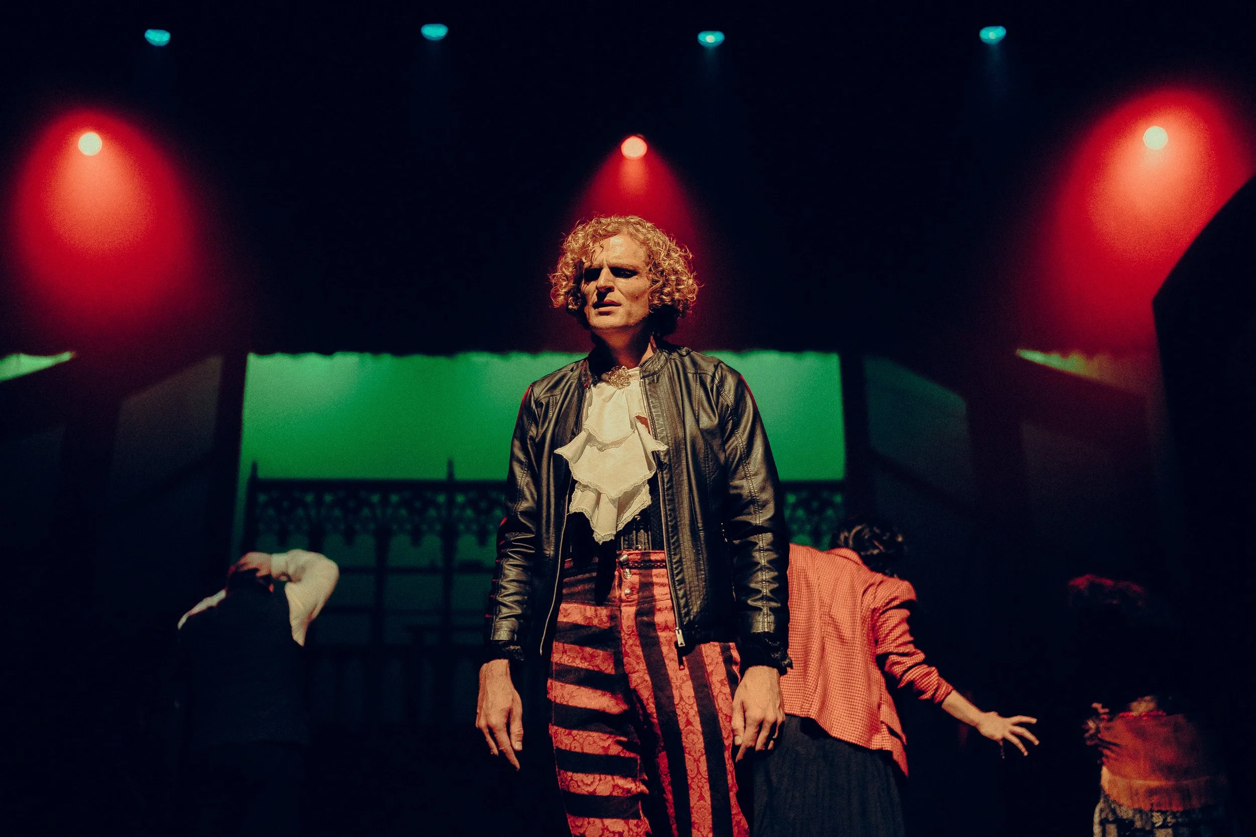A theatrical performance with a person in the foreground wearing a black leather jacket, ruffled shirt, and striped pants under dramatic stage lighting.