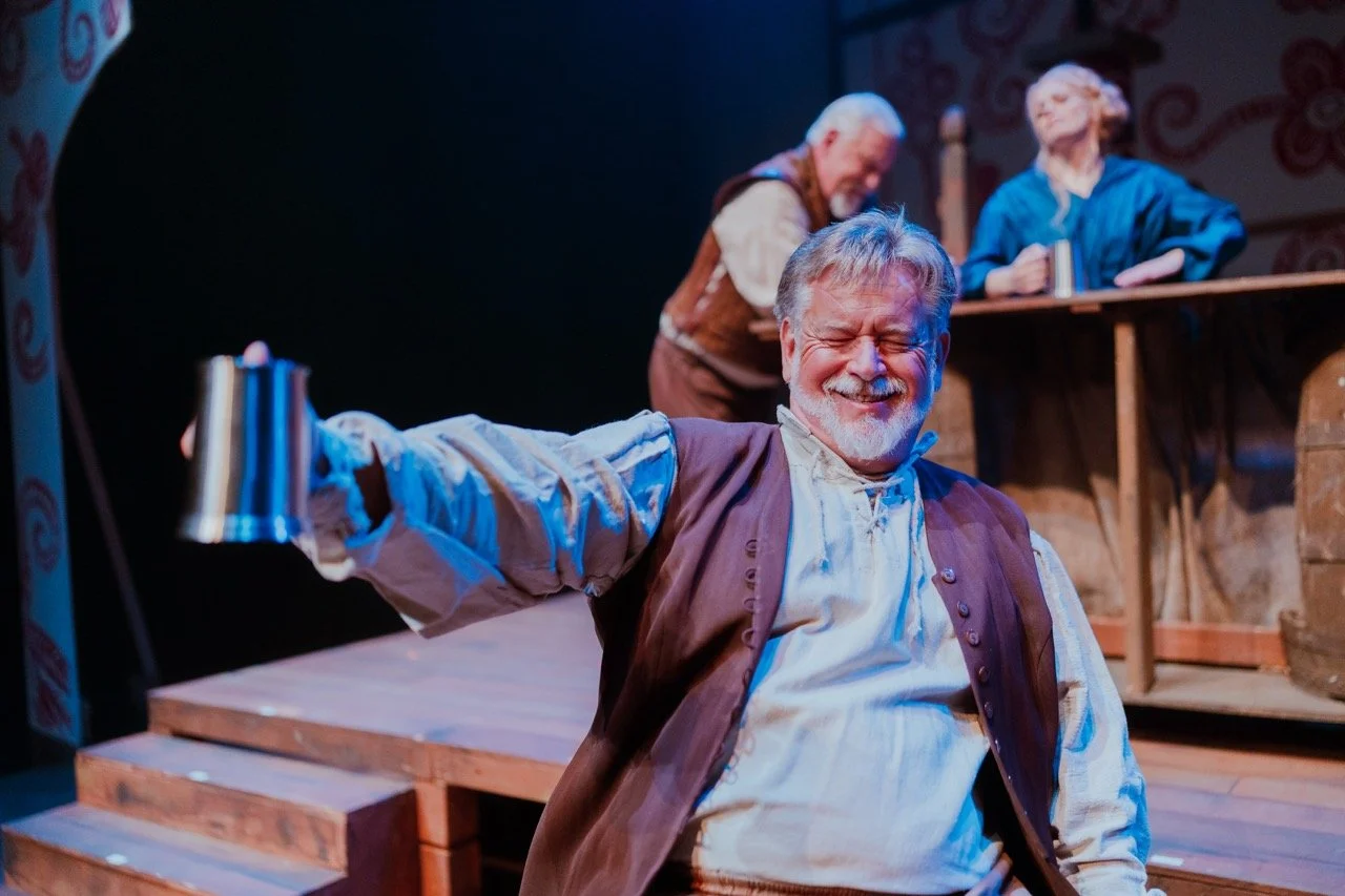 Actor in costume holding a metal mug during a stage performance with two other actors in the background near a bar.