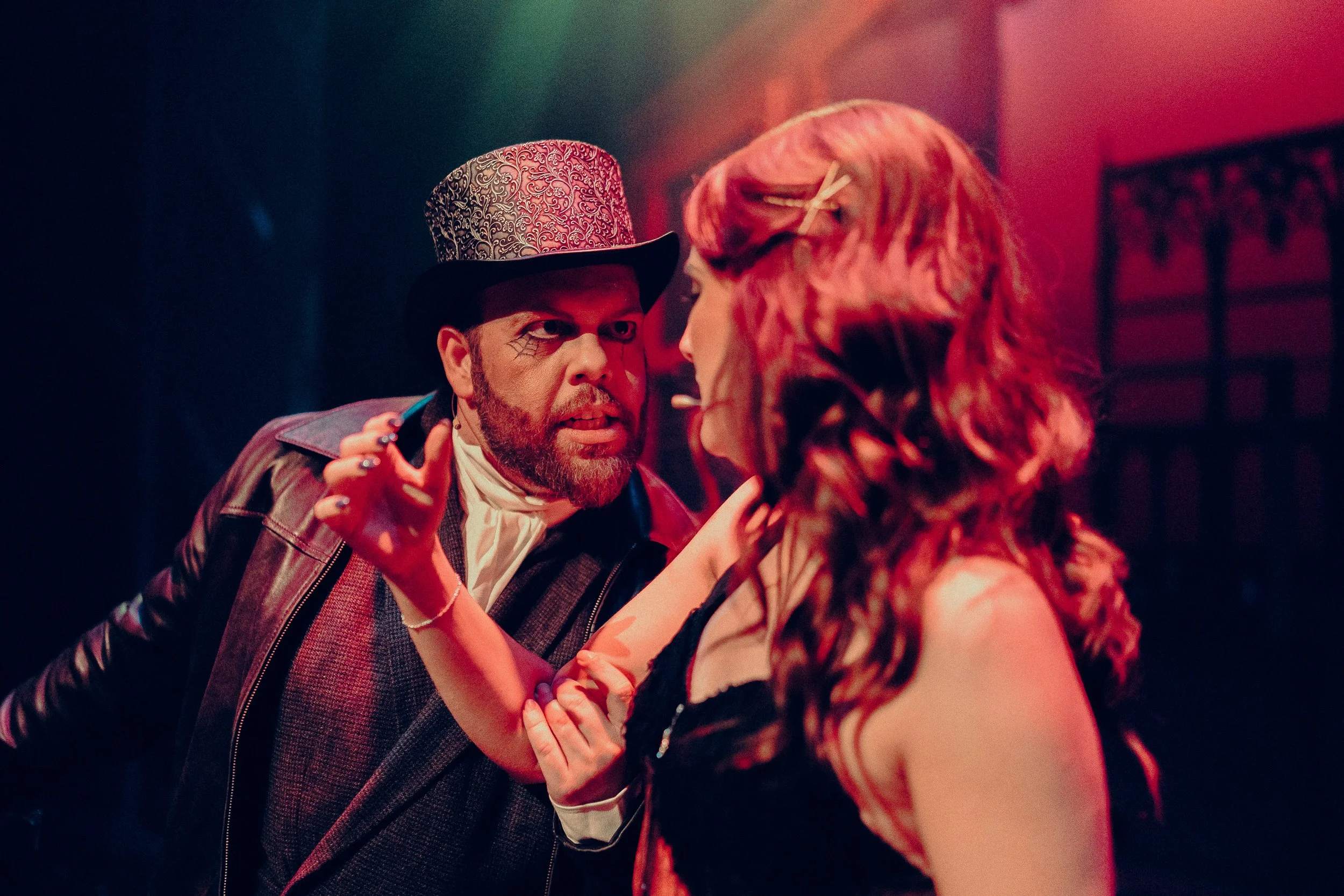 Two actors in dramatic stage lighting, one with a top hat and intense expression, the other with long hair, engaging in a theatrical scene.