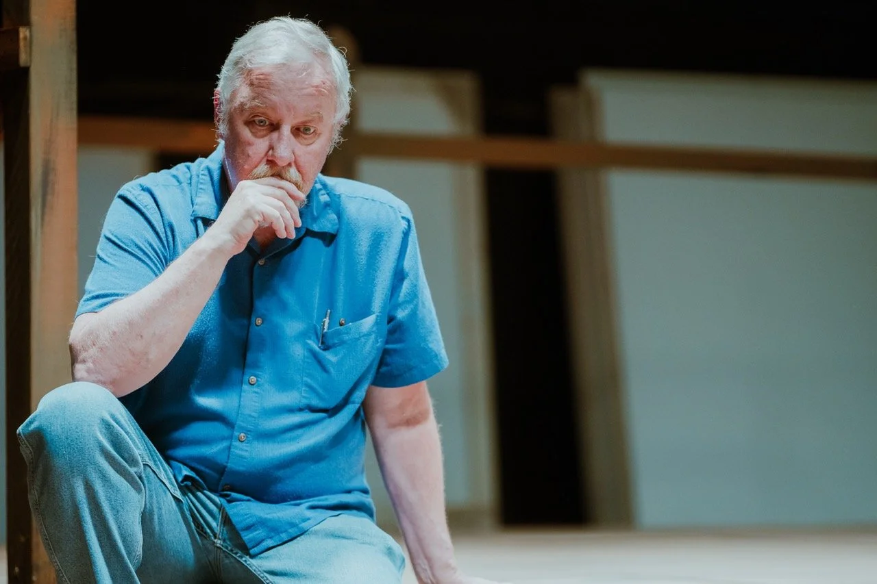 Elderly man in a blue shirt sitting pensively, with a thoughtful expression.