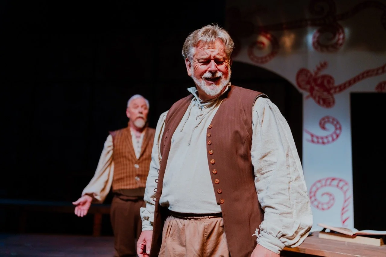 Two actors in period costume performing on stage, in a dramatic scene with expressive gestures, one in the foreground and one blurred in the background, with decorative backdrop.