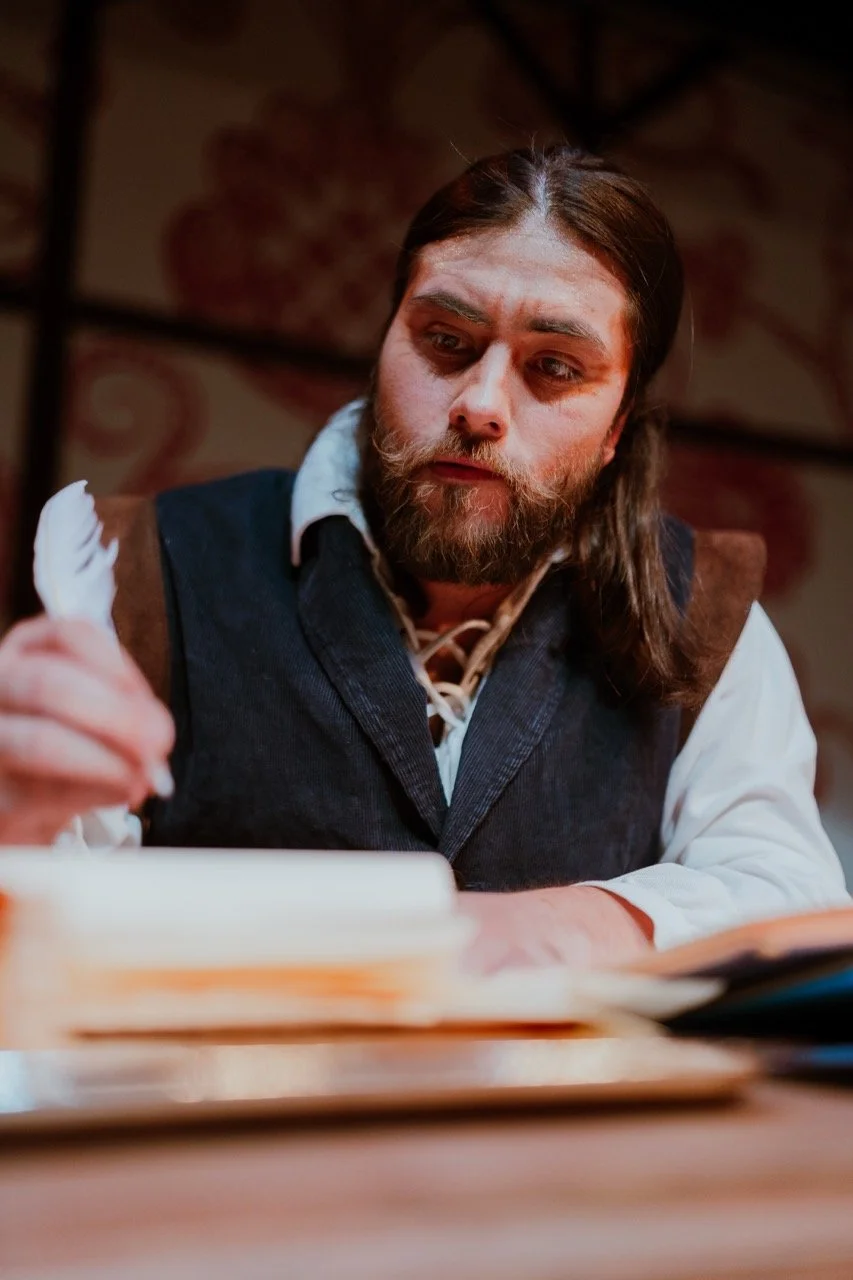 Man with long hair and beard writing with quill pen in period clothing.