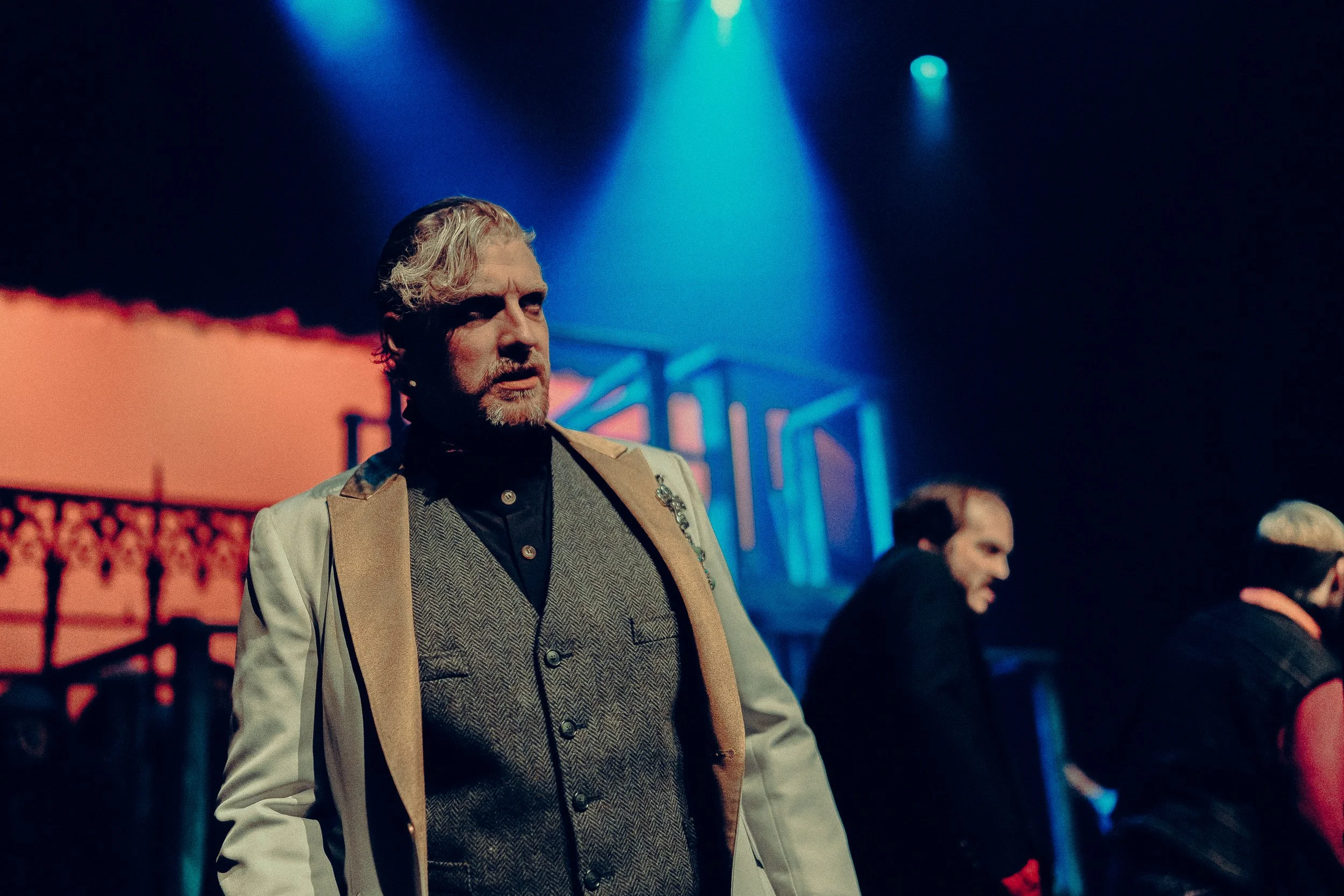 Theatrical performance with dim lighting; a man in a white jacket and gray waistcoat in focus, with background characters and stage set.
