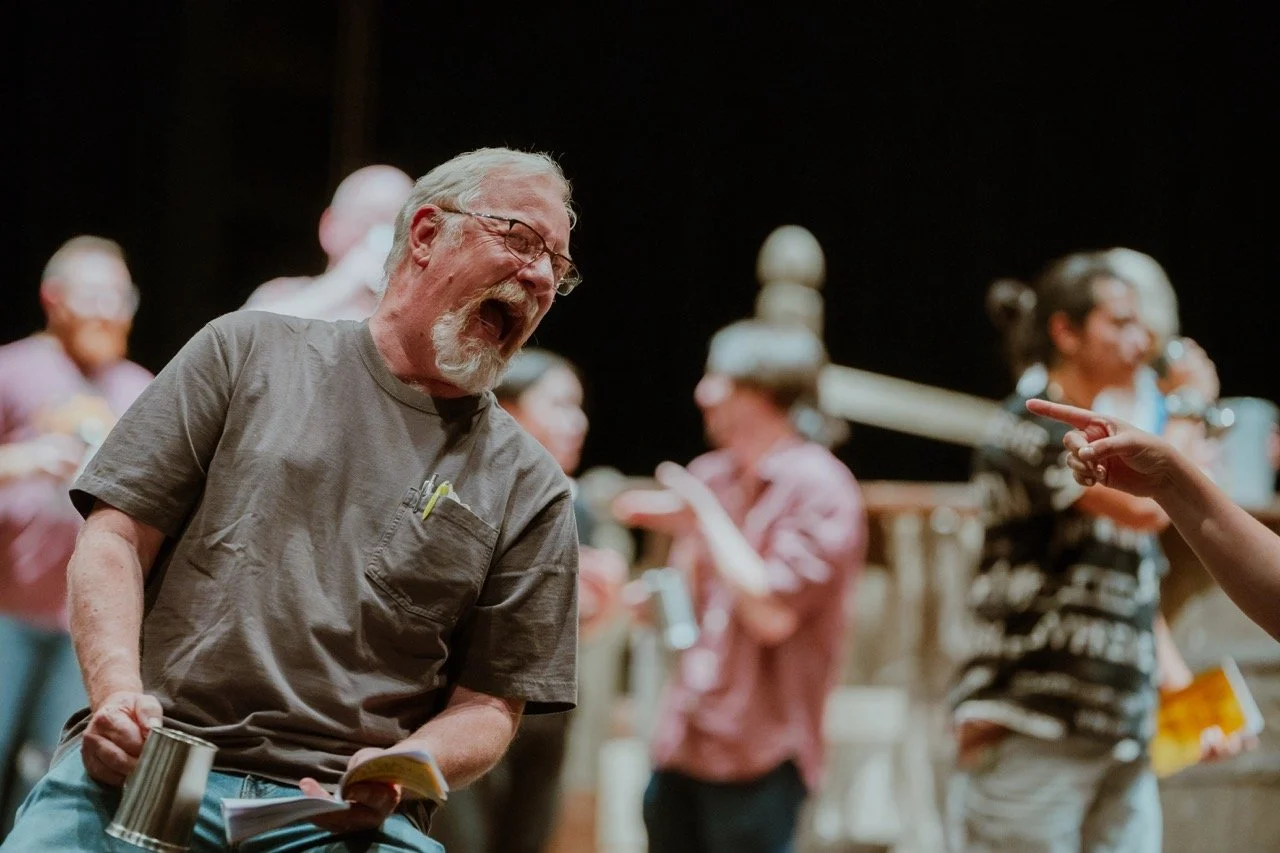 A group of people engaged in a lively discussion or rehearsal on stage, with one person in the foreground appearing animated and holding a mug and a script. Others in the background are interacting or observing.
