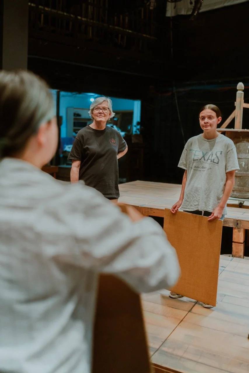 Three people in a theater setting, one in focus holding a piece of cardboard, two others conversing. Background is dimly lit.