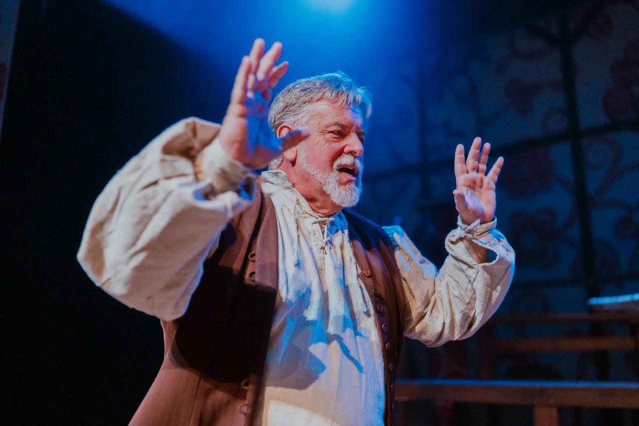 An older man with a gray beard wearing a historical costume gestures with his hands on stage under blue lighting.