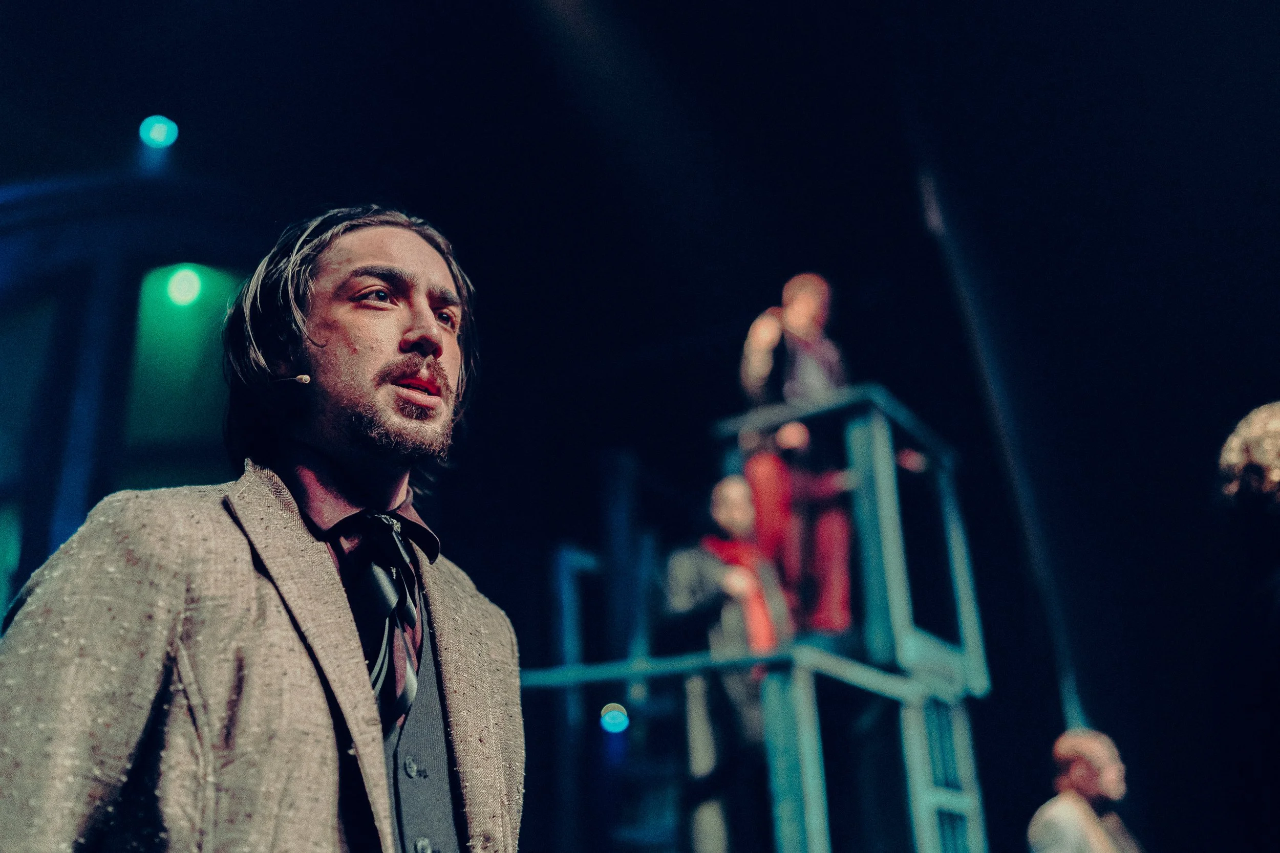 A man in a theatrical performance wearing a microphone headset and a suit, with two other people in the blurred background standing on a platform.