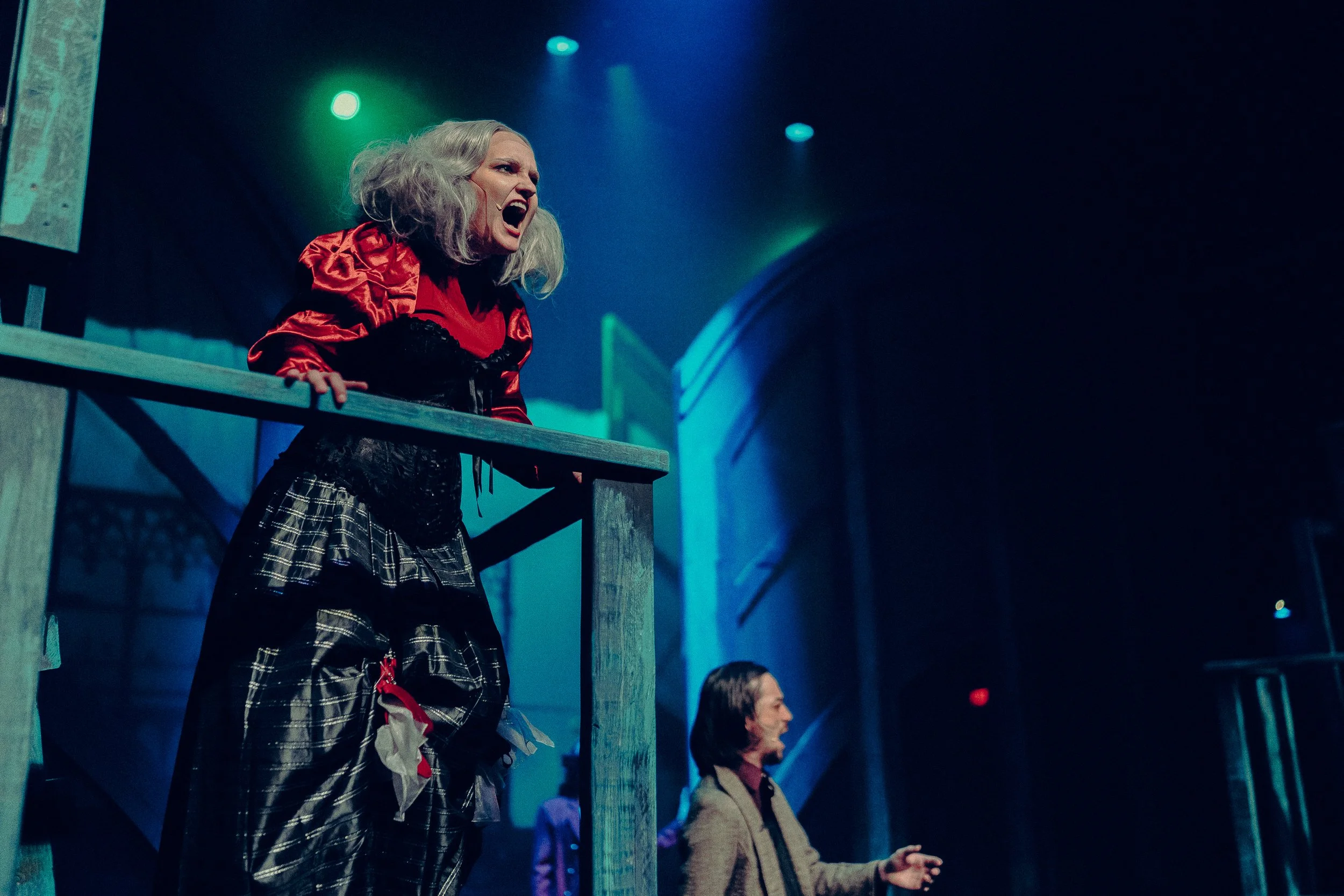 Two actors performing on stage, with one wearing a dramatic costume and expressing intense emotion, and the other in the background. The setting is dimly lit with blue and green stage lights, giving a theatrical ambiance.