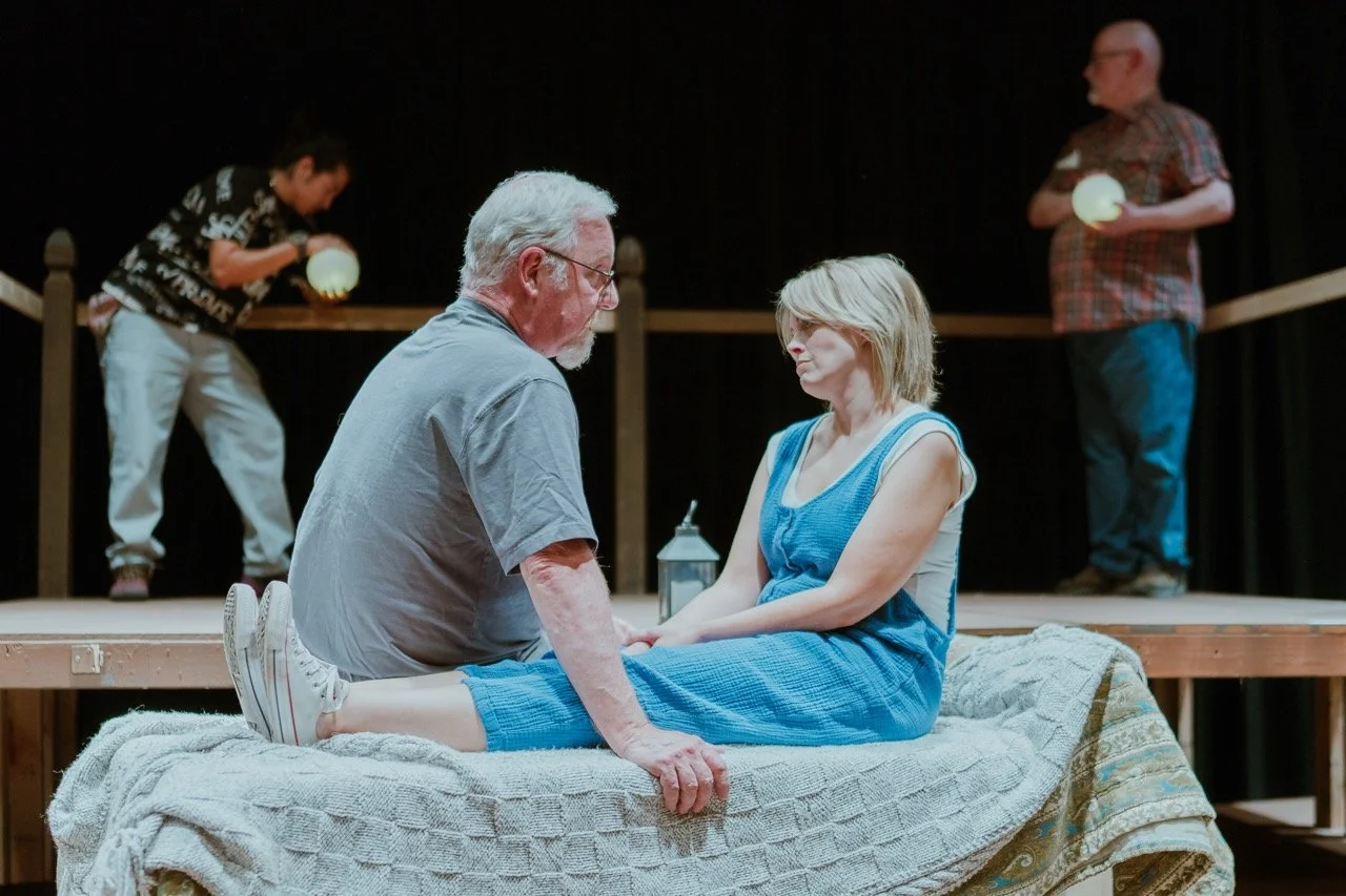 Four people on stage, two seated on a cushioned platform having an intense conversation, while two others stand in the background holding spherical objects.