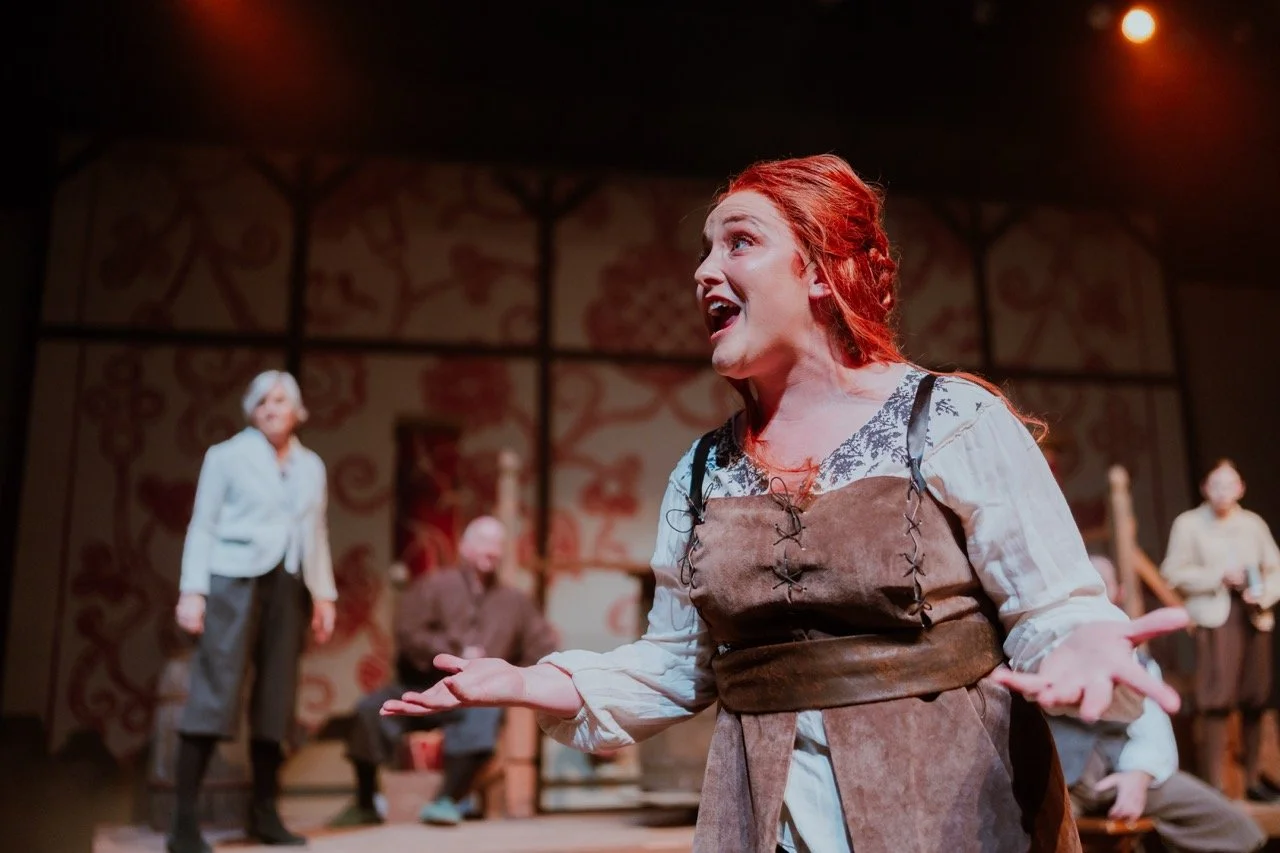 Theatrical performance with actors on stage, featuring a woman in a medieval-style costume with her arms outstretched, expressing emotion. Background includes other actors and a patterned backdrop.