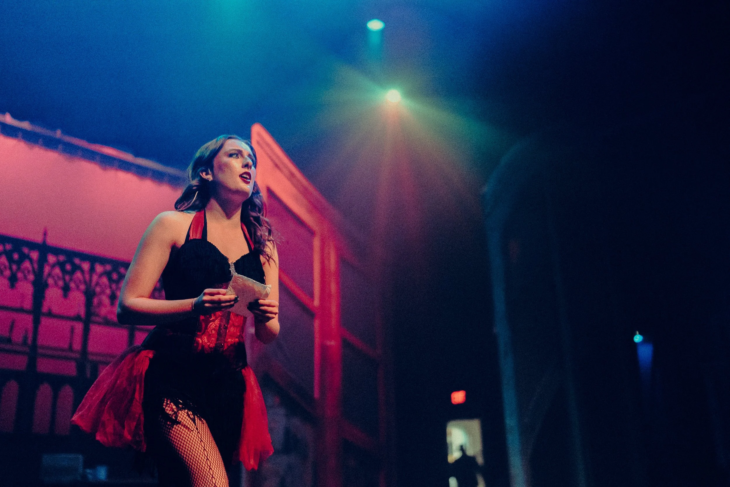 A performer on stage wearing a black and red costume, holding a piece of paper, with dramatic lighting in a theater setting.