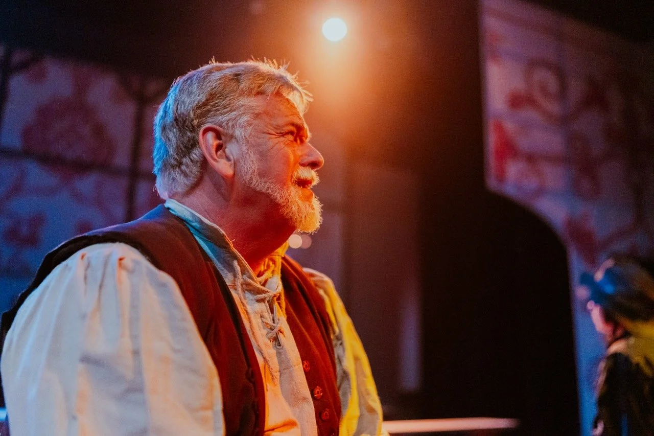 A man in historical costume, including a vest and shirt, on stage with dramatic lighting.