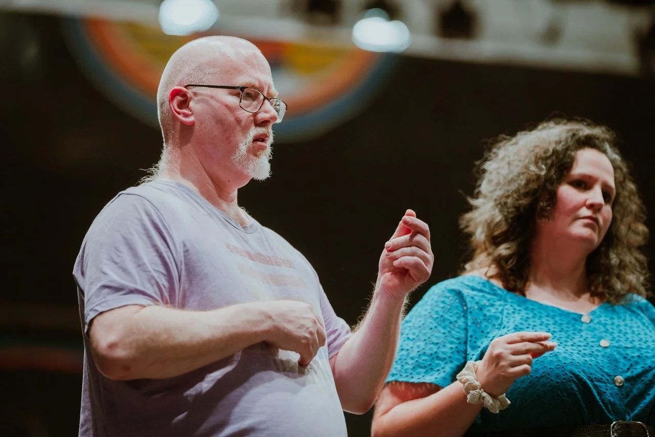 Two individuals, one bald with glasses and the other with curly hair, standing indoors, gesturing with their hands. The bald person is wearing a light purple shirt, and the person with curly hair is wearing a blue top.