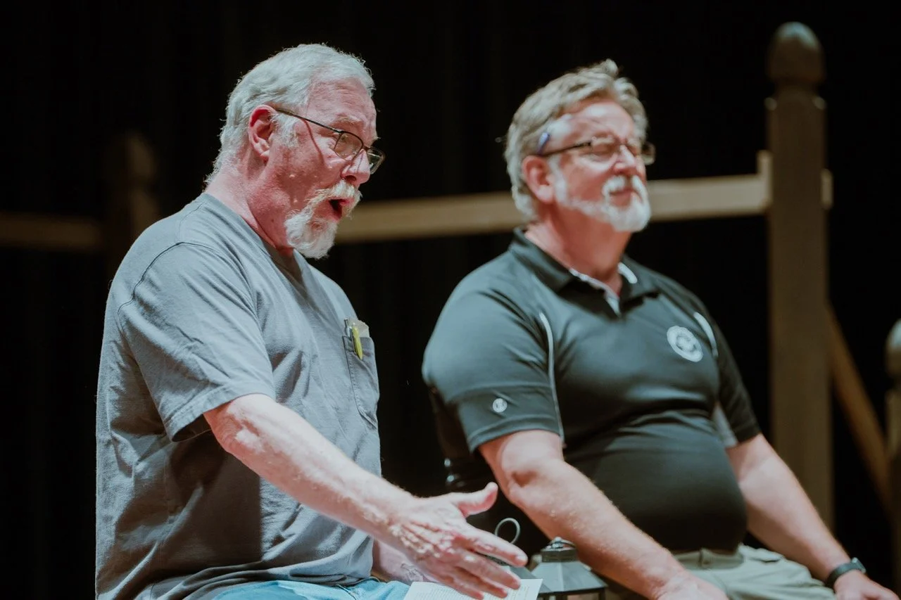 Two older men sitting on stage, one speaking passionately, with papers in hand, and the other listening attentively, in a dimly lit setting.