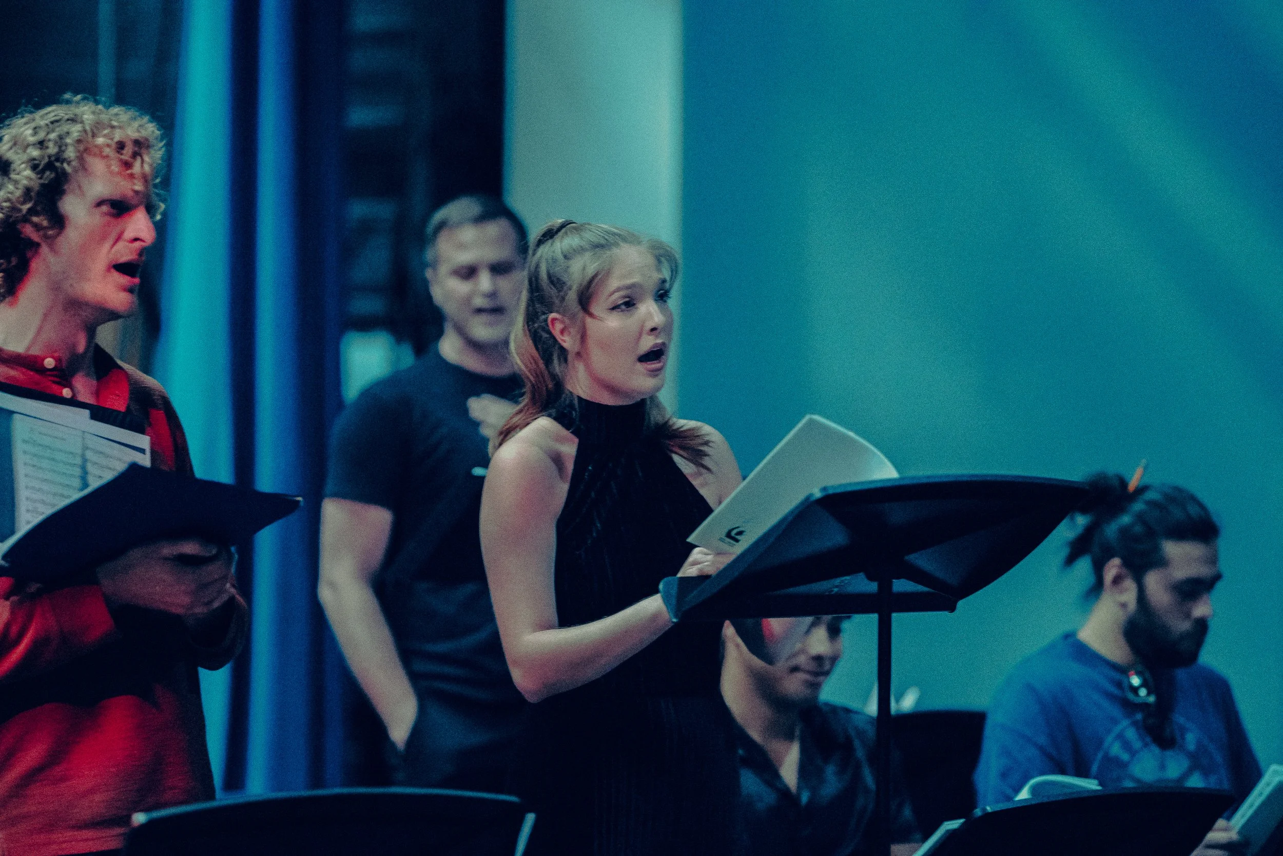 A group of singers rehearsing in a music studio, with a woman in the foreground holding sheet music and singing.