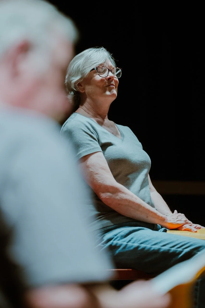 Elderly woman in glasses seated with hands in lap, focus on background, soft focus on person in foreground.