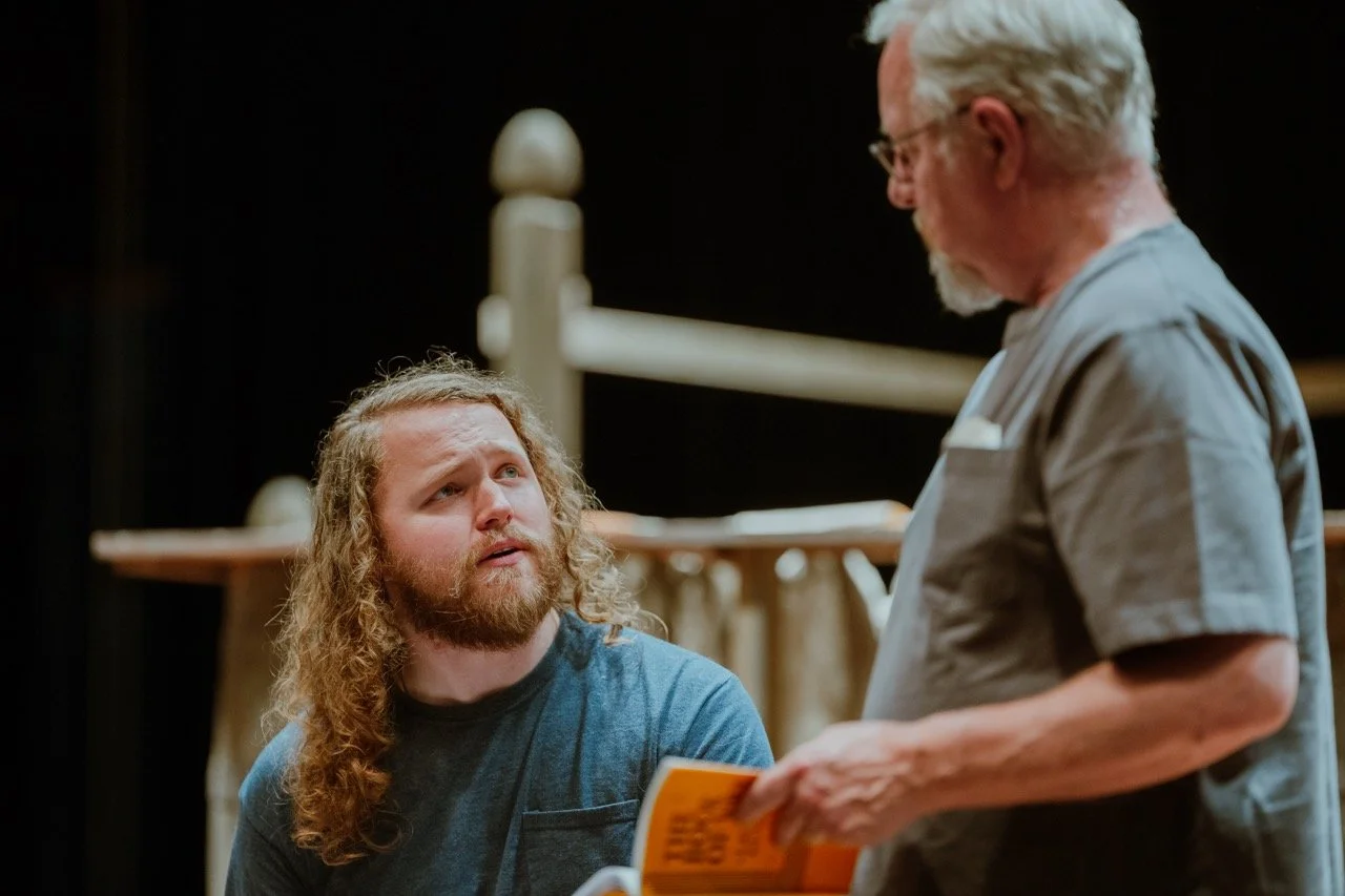 Two men interacting, one is holding a book while the other is sitting, with a wooden structure in the background.