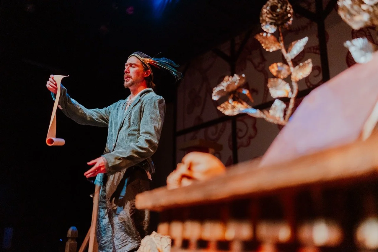 An actor in medieval costume holds a parchment scroll while performing on stage. The set includes a metallic rose and a skull, suggesting a theatrical or Shakespearean theme.