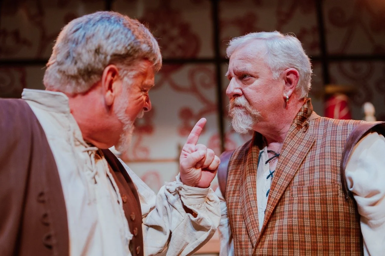 Two elderly men with white hair and beards are engaged in an intense conversation. One man is pointing his finger at the other, appearing to be in a heated discussion. They are both wearing old-fashioned, rustic clothing, possibly indicating a theatr