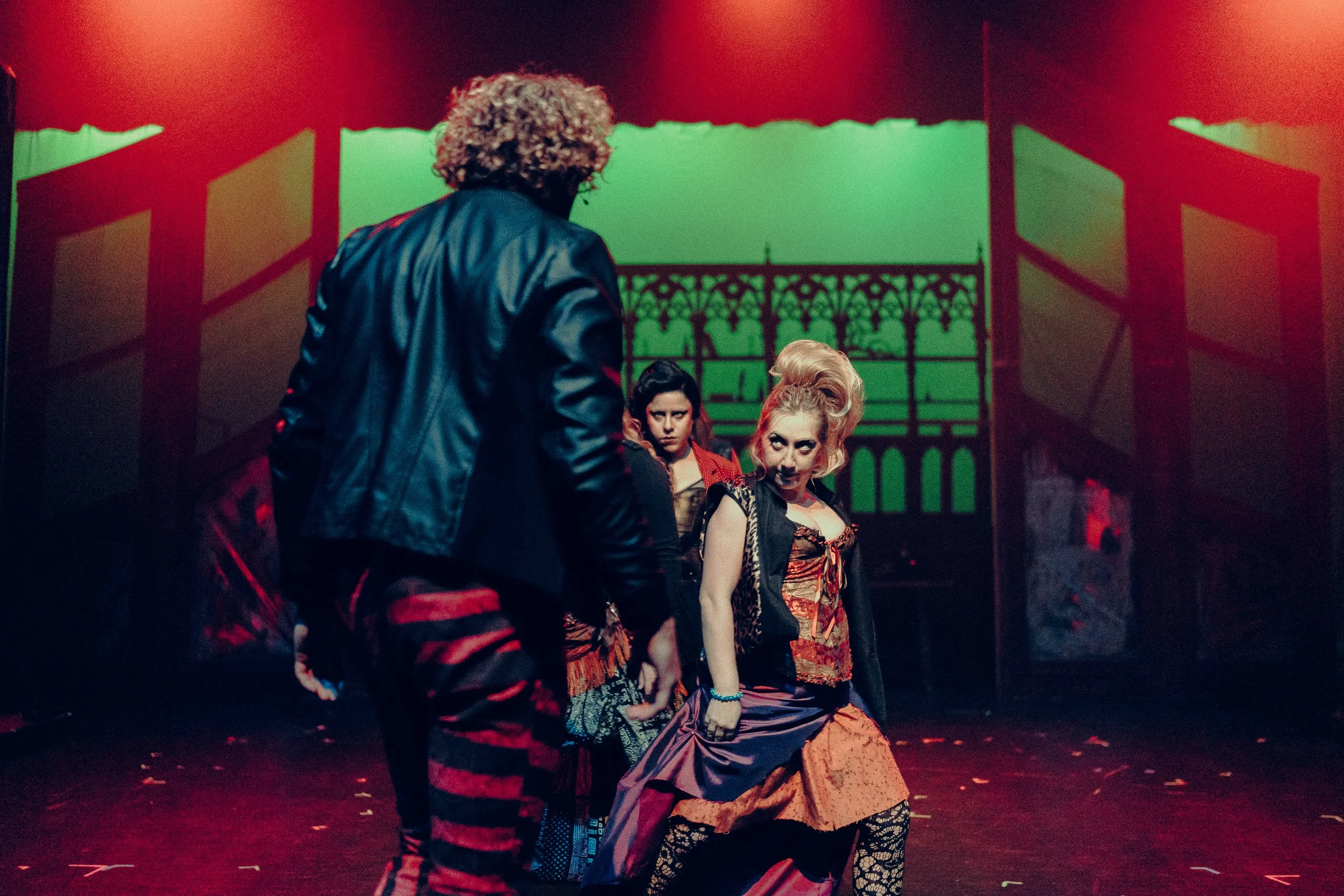 Theater performance with three actors in colorful, Gothic costumes against a dimly lit stage backdrop.