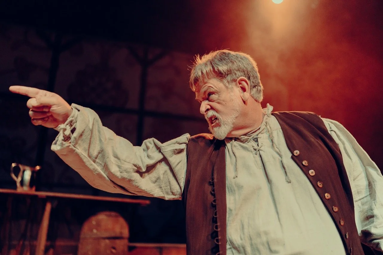 A man in period clothing pointing emphatically during a stage performance, with a dramatic expression under warm stage lighting.
