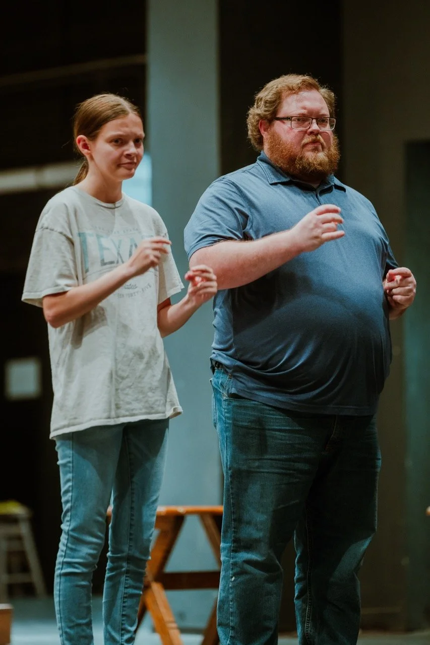 Two people standing side by side in a casual indoor setting, both wearing jeans and T-shirts. The person on the left is young and wearing a light-colored top. The person on the right has a beard and is wearing a blue polo shirt. They appear to be in