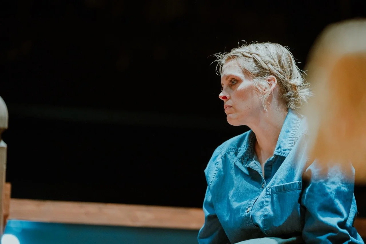 A person wearing a denim shirt, looking pensive, against a dark background.