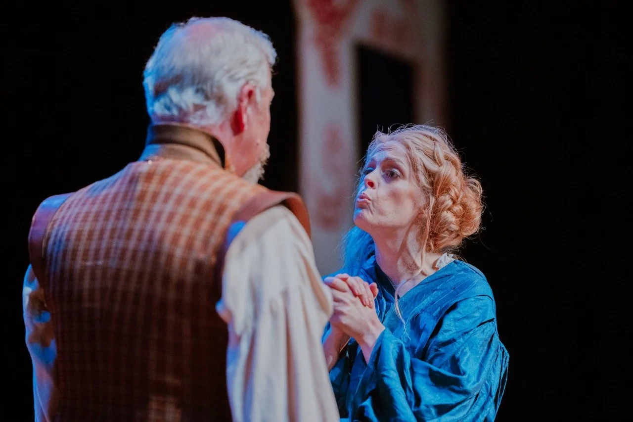 Two actors in period costumes performing on stage, one man in a checked vest and one woman in a blue dress, appearing in an emotional scene.