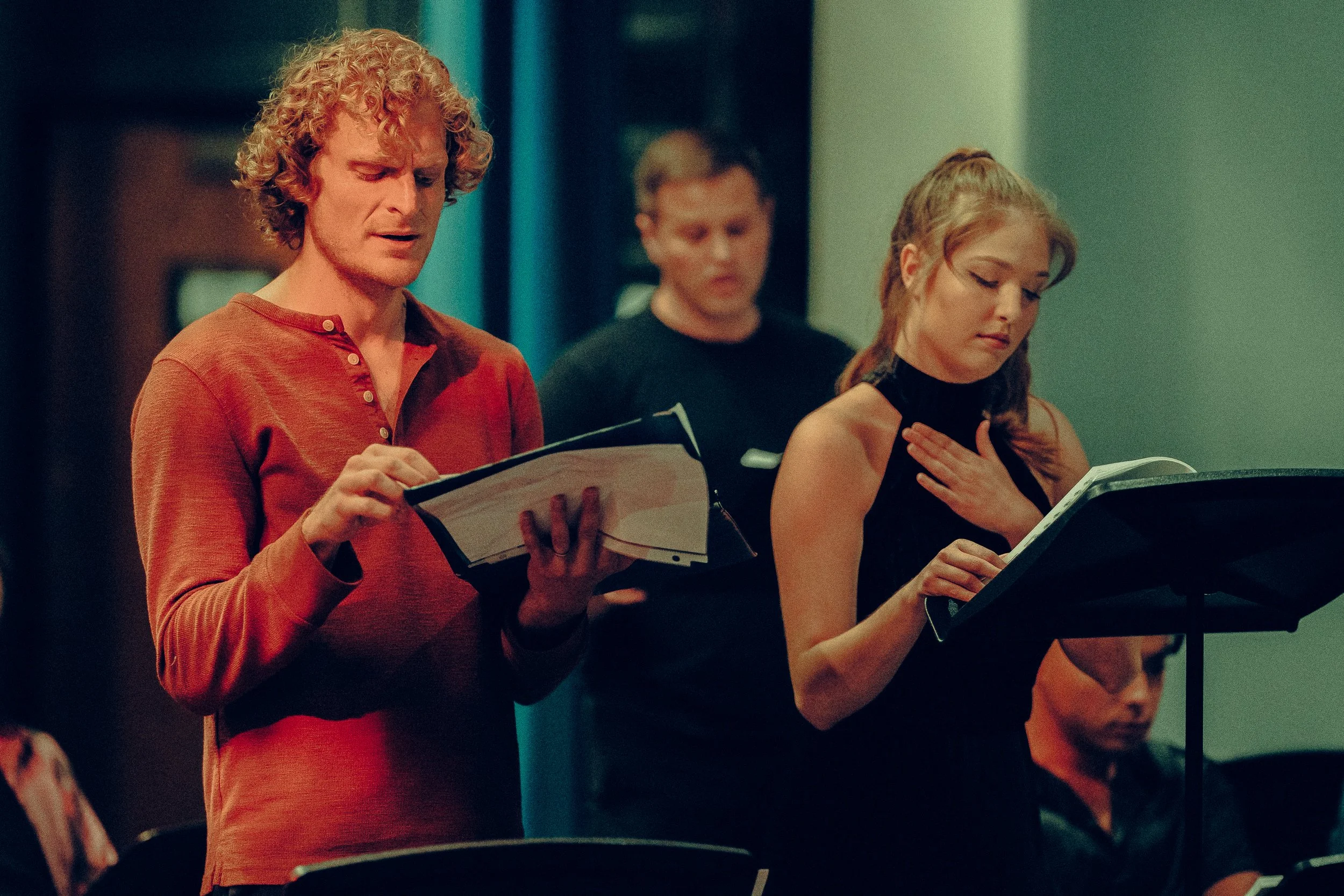 People in a rehearsal, two holding scripts, one reading, the other gesturing with hand on chest, others in background.