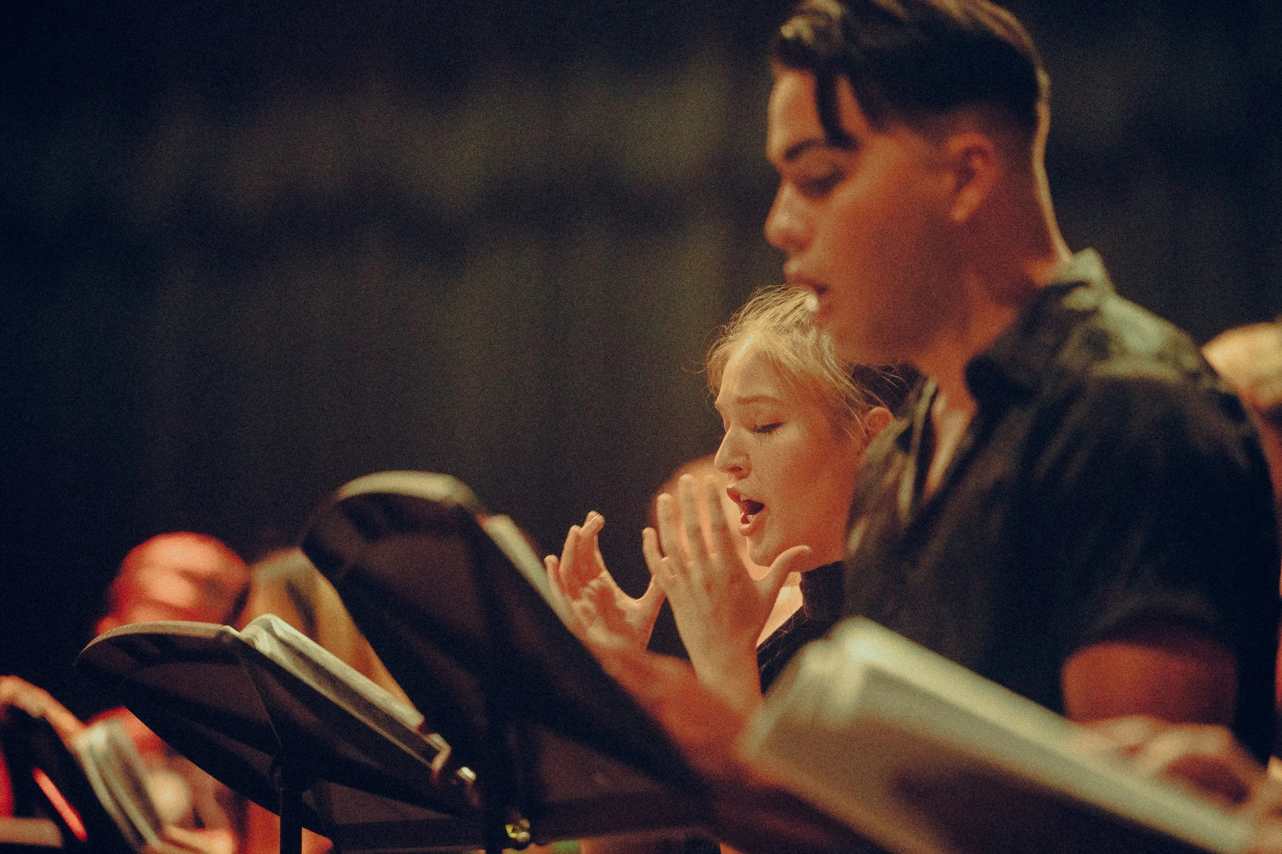People singing in a choir, holding sheet music, with focused expressions in a dimly lit setting.