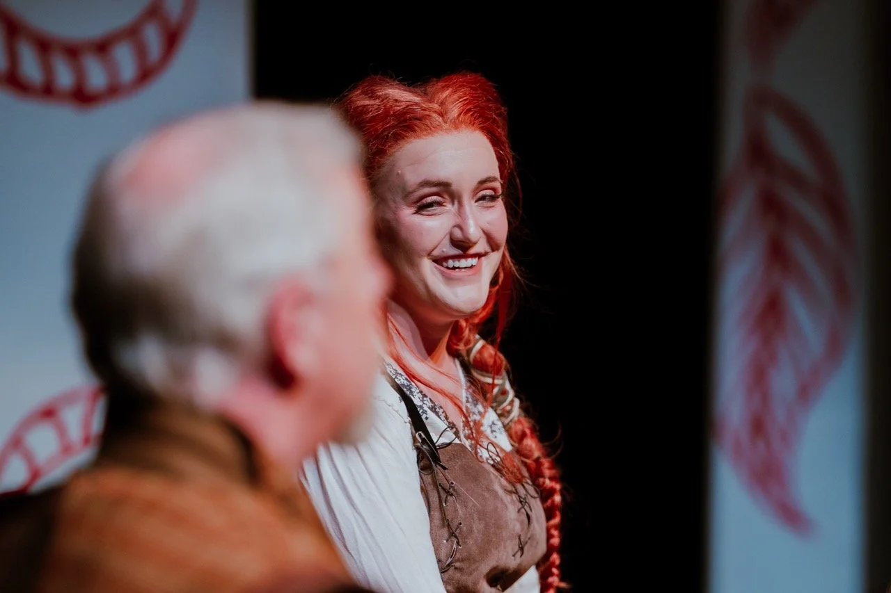 A woman with red hair smiling on stage during a performance, wearing a costume with a white blouse and brown vest. Blurred person in the foreground.