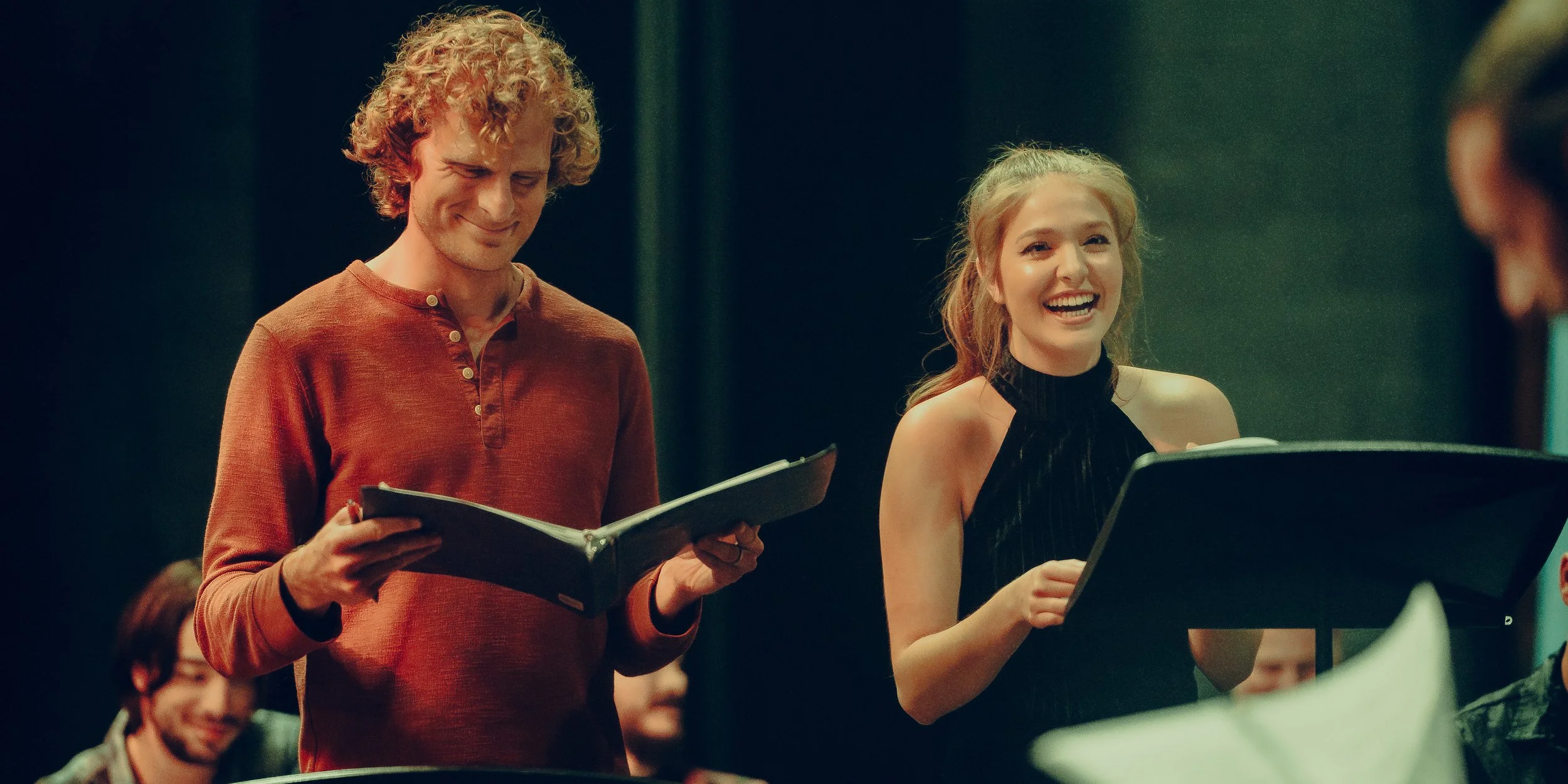 Two people reading scripts on stage, smiling and engaged, possibly rehearsing for a performance.