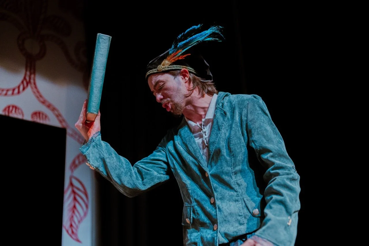 Actor in costume performing on stage, wearing a blue coat and feathered hat, holding a book enthusiastically during a theatrical play.