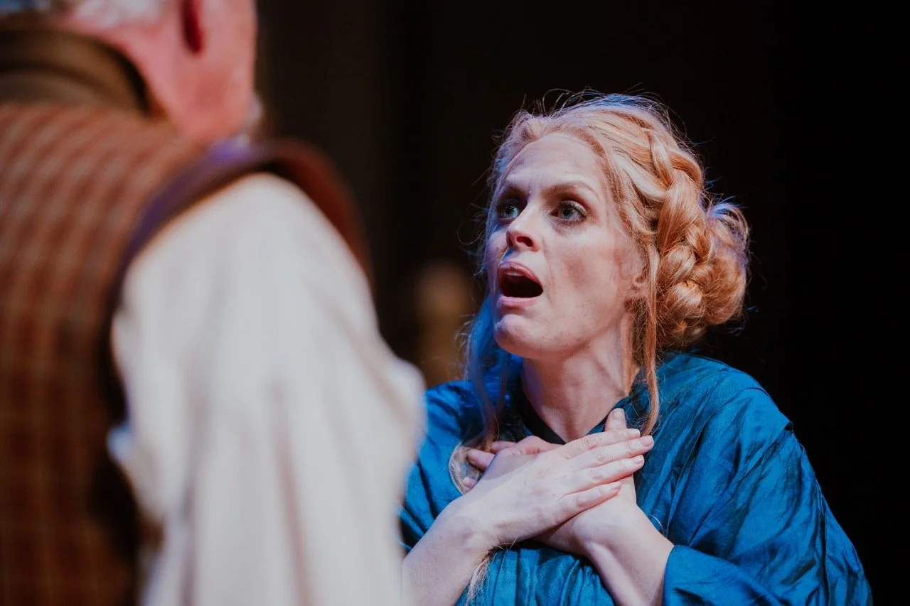 A woman with blonde hair in an updo, wearing a blue blouse, appears emotional with hands clasped to her chest, facing a man in a plaid vest, in a theatrical scene.