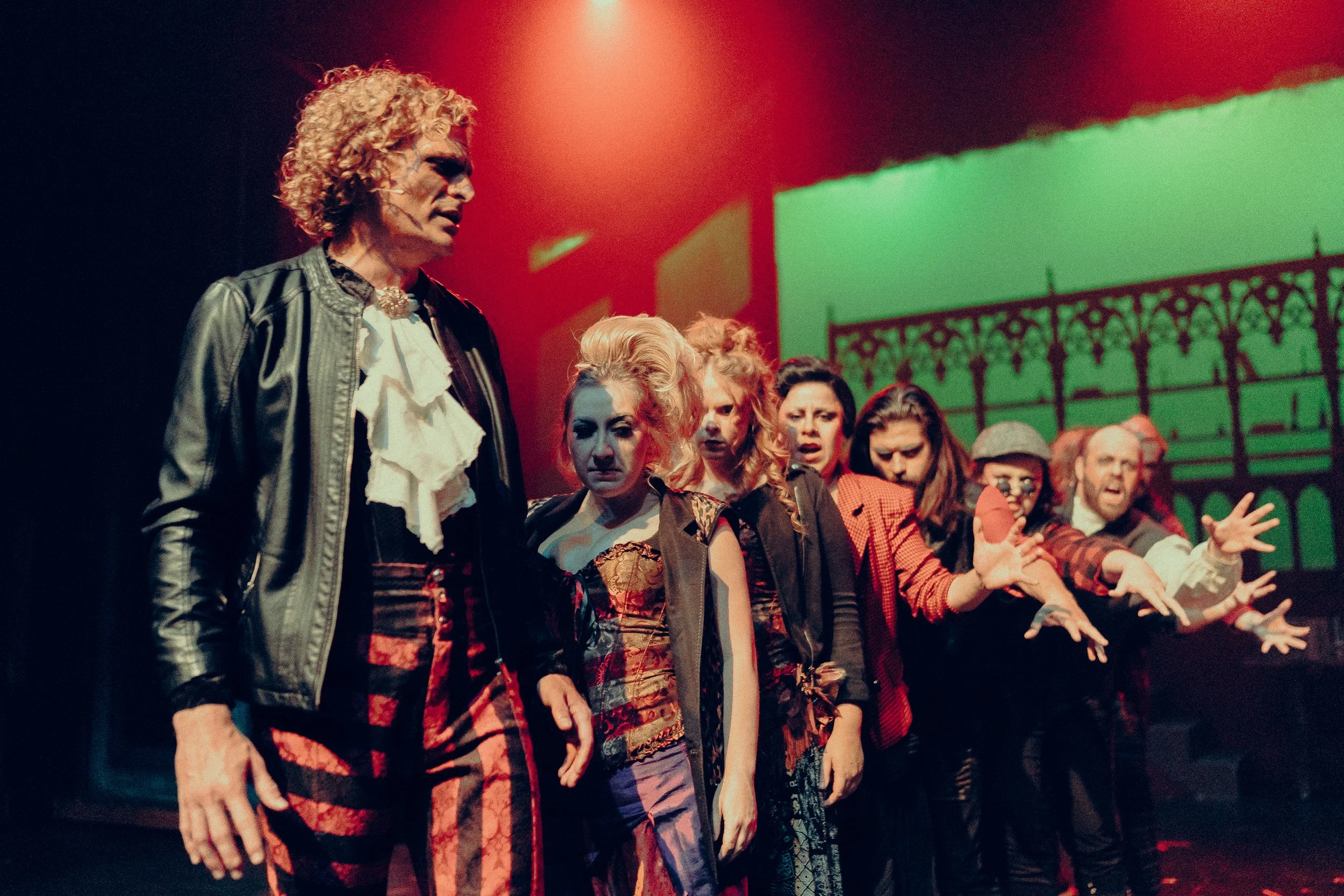 Theatrical performance with actors in costume under dramatic lighting, including one with curly hair in a black leather jacket and others in ornate, colorful clothing, standing in a line.