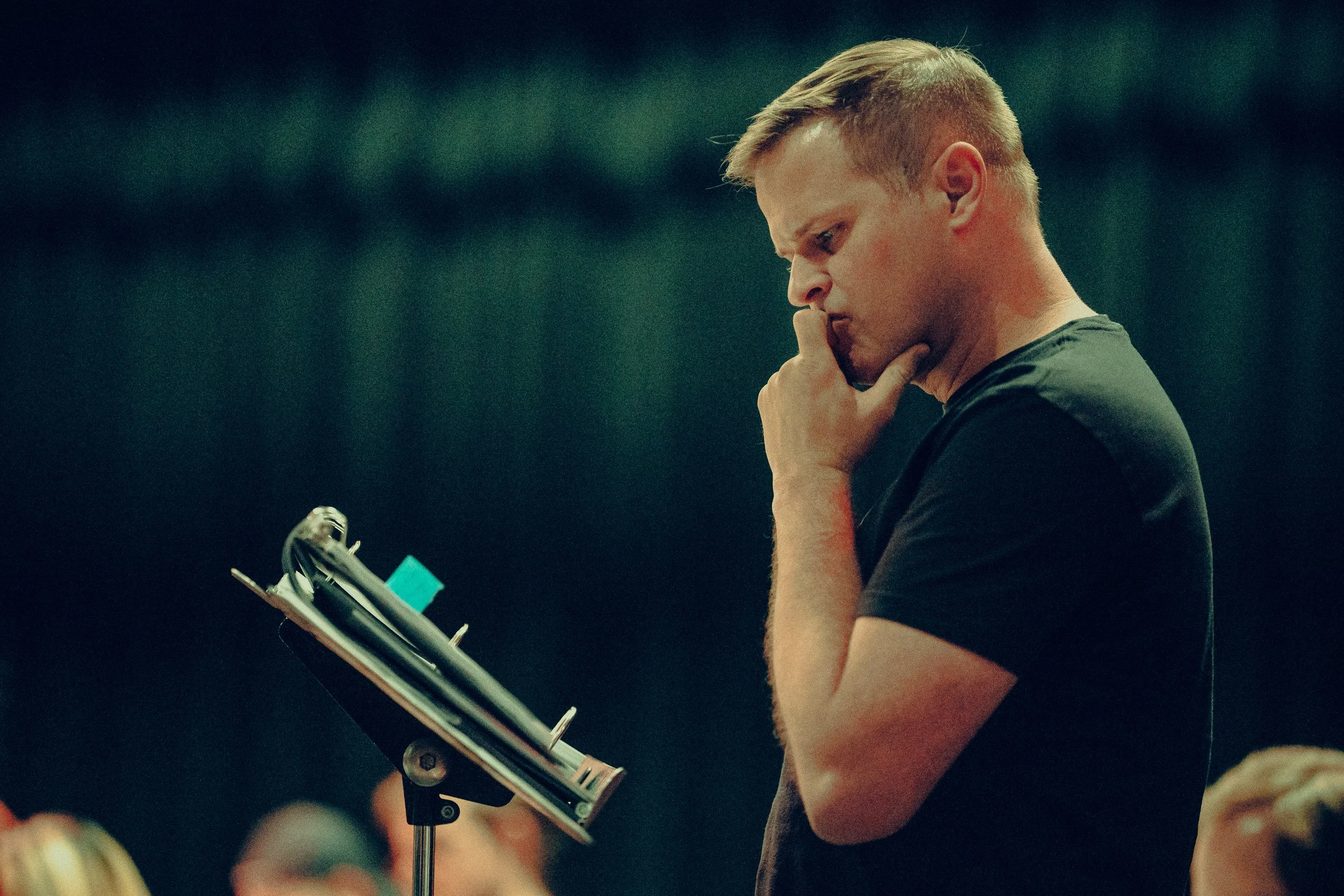 Man in a black shirt standing thoughtfully in front of a music stand, holding his chin, with a blurred background.