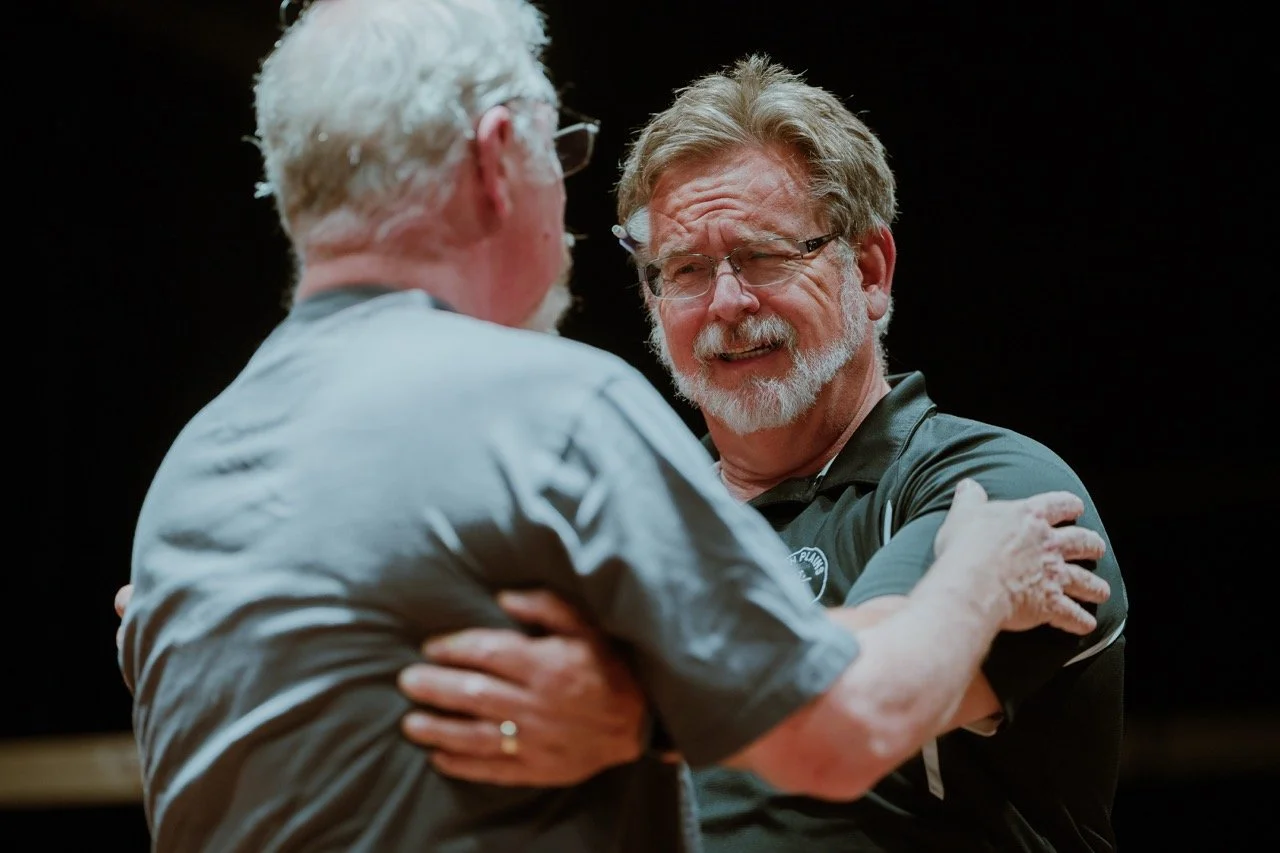 Two older men with beards embracing and smiling, wearing casual clothes, in a dark setting.
