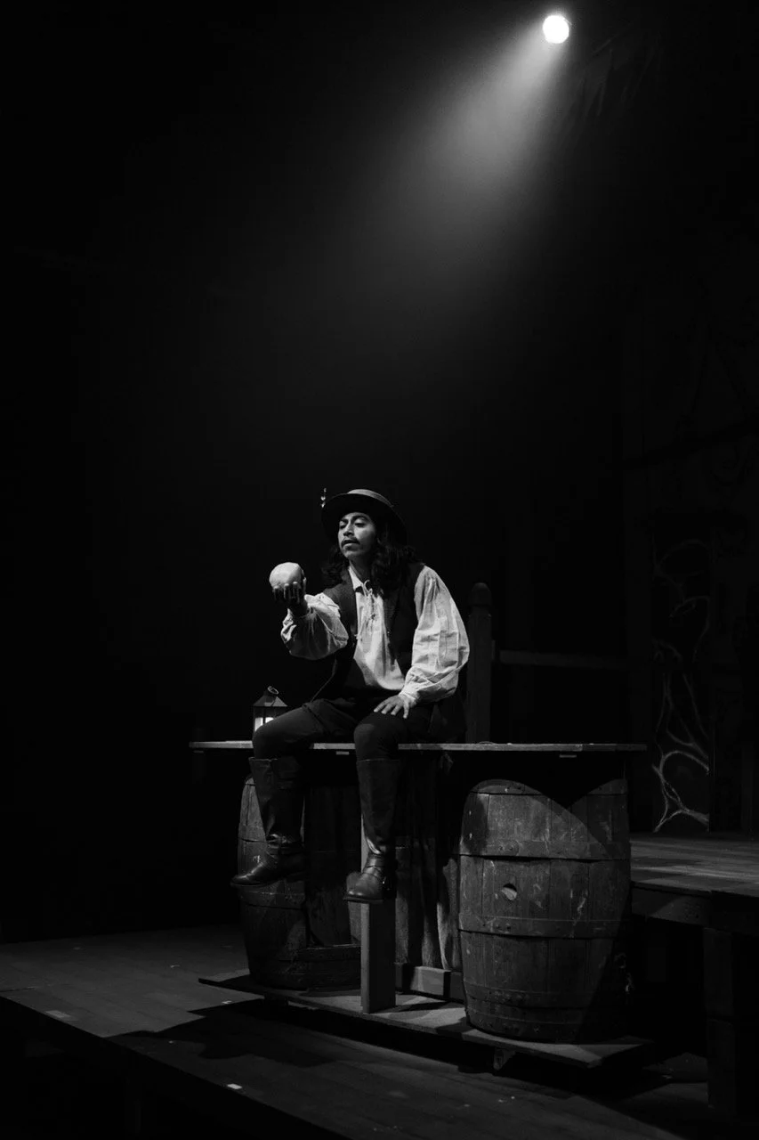 Black and white photo of a theatrical performance featuring a person in a historical costume holding a skull, sitting on wooden barrels on stage, illuminated by a spotlight.