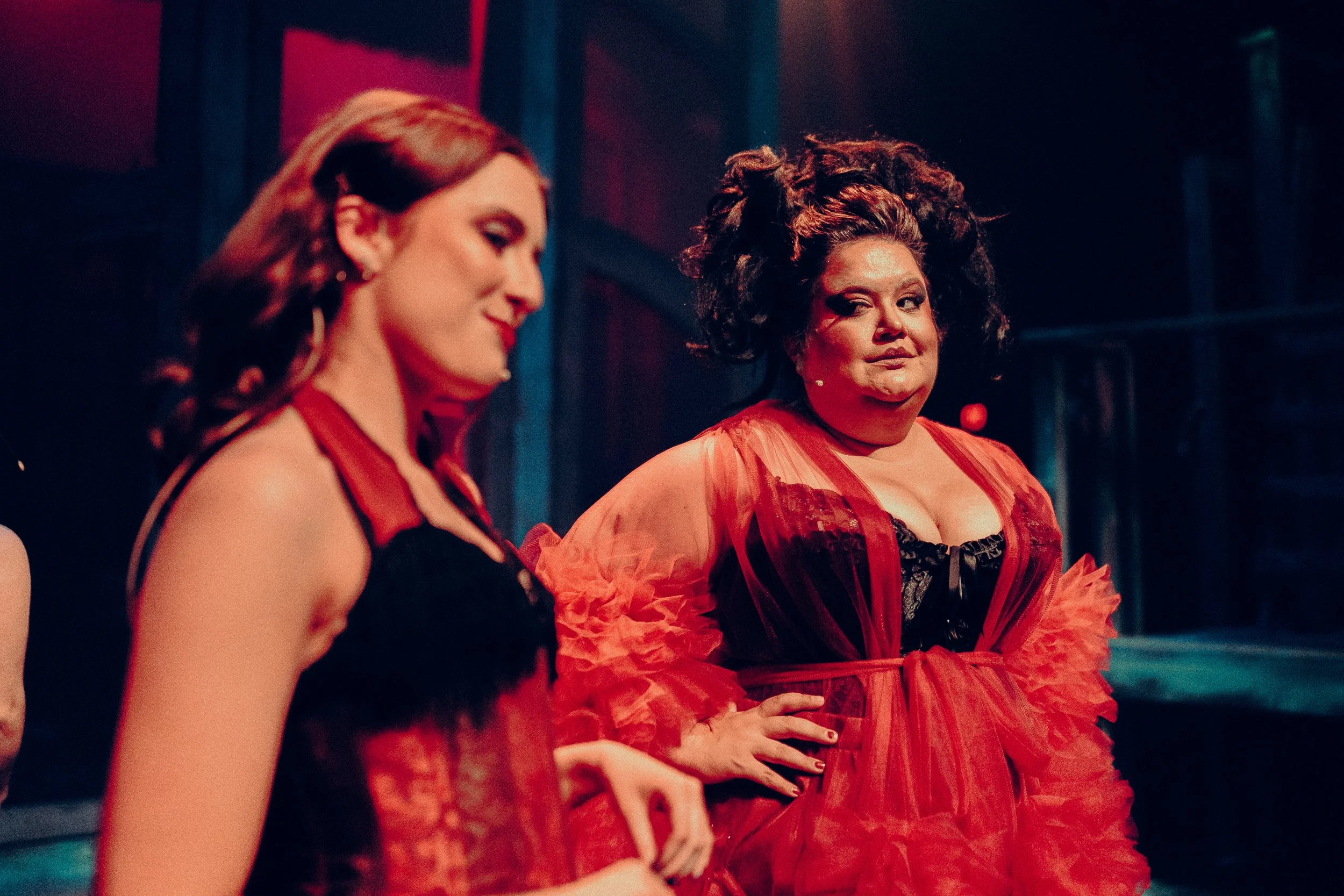 Two performers in a theater setting wearing dramatic costumes with bright stage lighting.