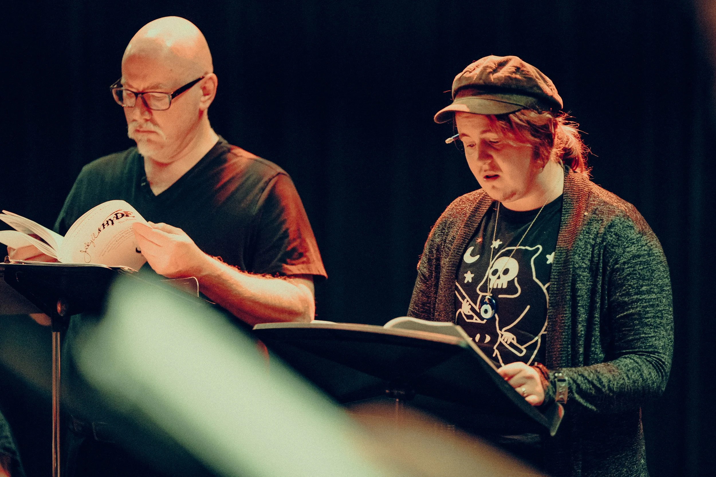 Two people reading scripts on stage in dim lighting, one wearing a T-shirt with a skull design