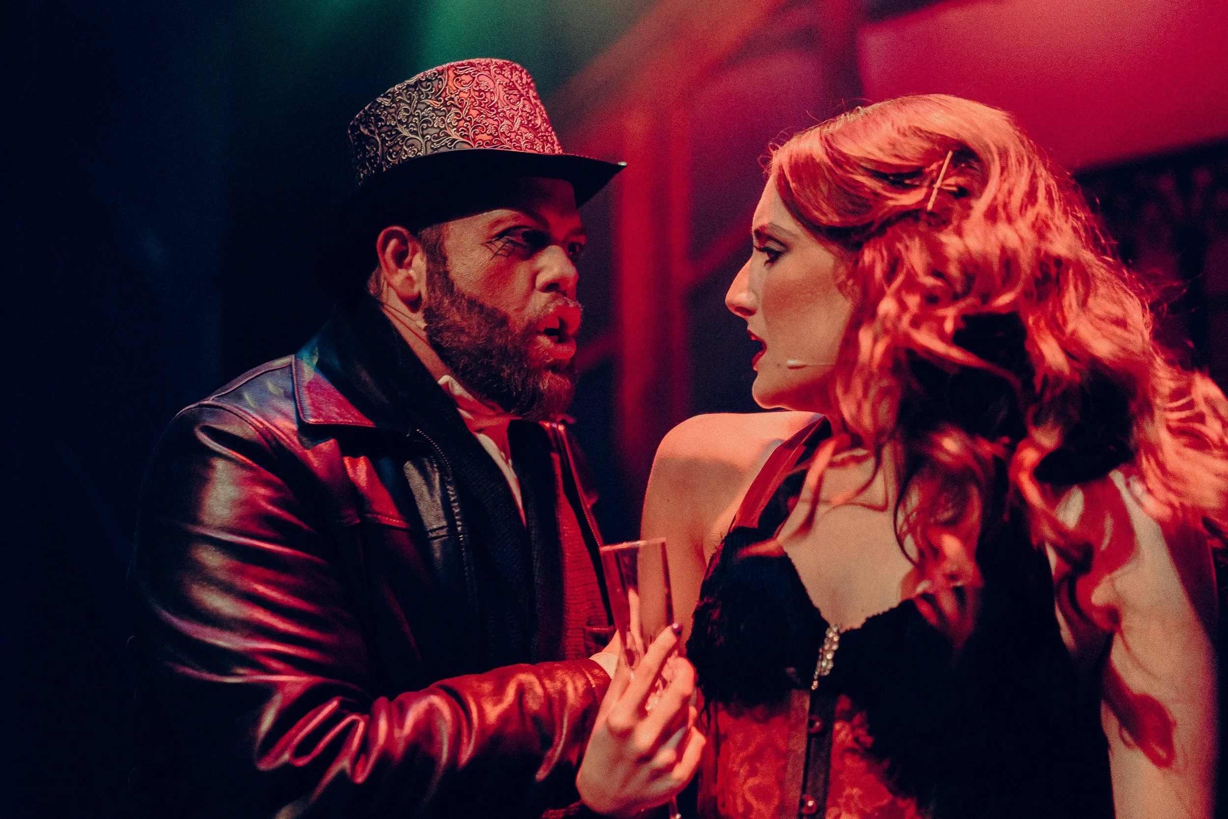 Two performers on stage in dramatic lighting, a man in a leather jacket and ornate top hat, and a woman in a red corset with long wavy hair.