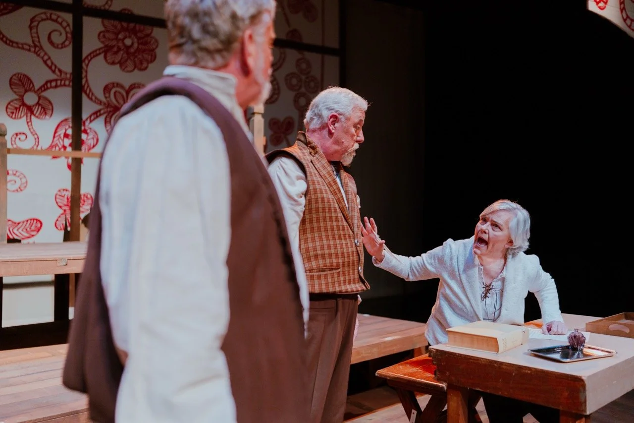 Three actors performing an emotional scene on stage, with two elderly men standing and one elderly woman seated at a table, gesturing passionately. The background features a decorative floral pattern.