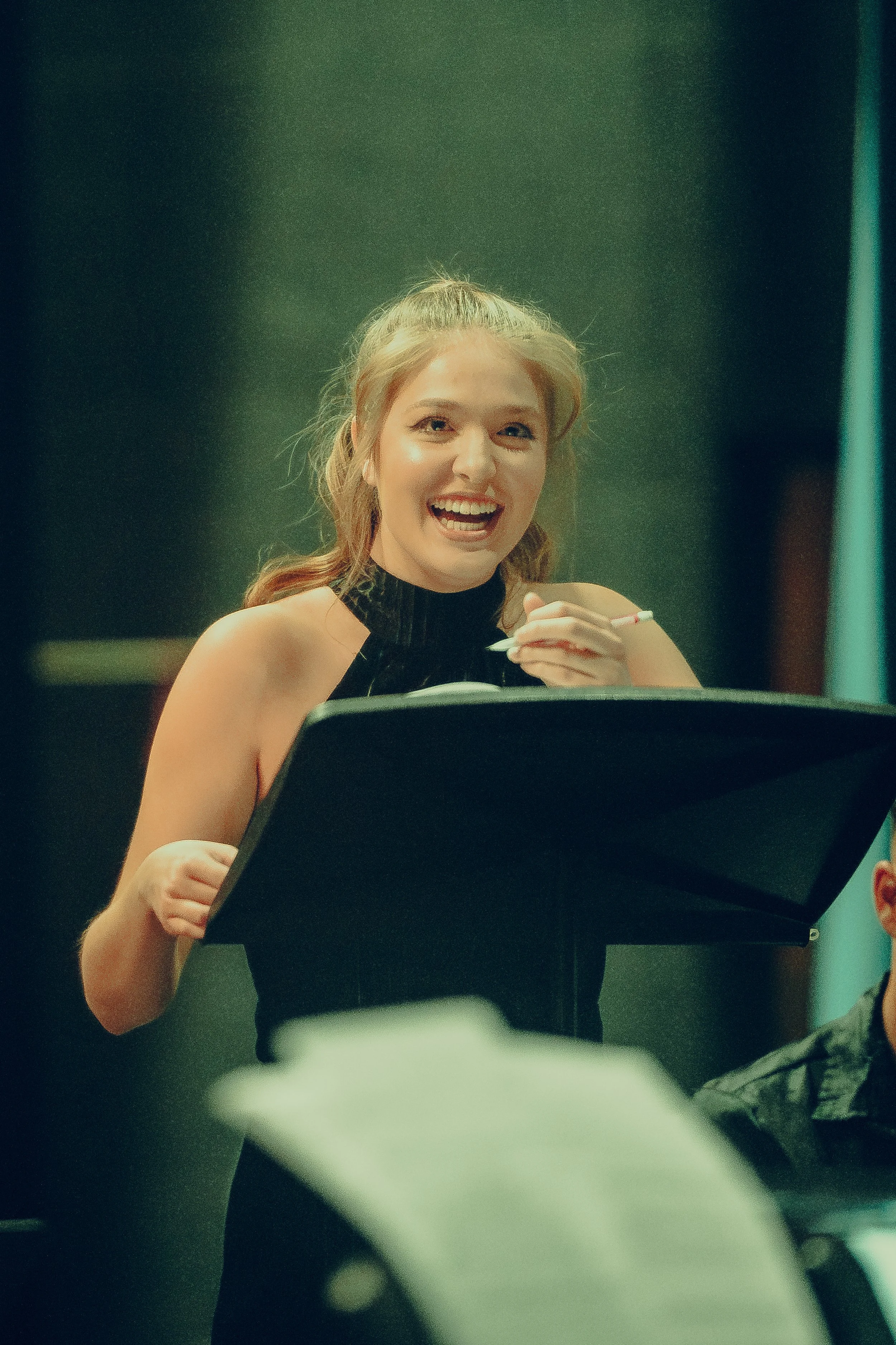Woman speaking at a podium, holding papers, with a joyful expression in a dimly lit setting.