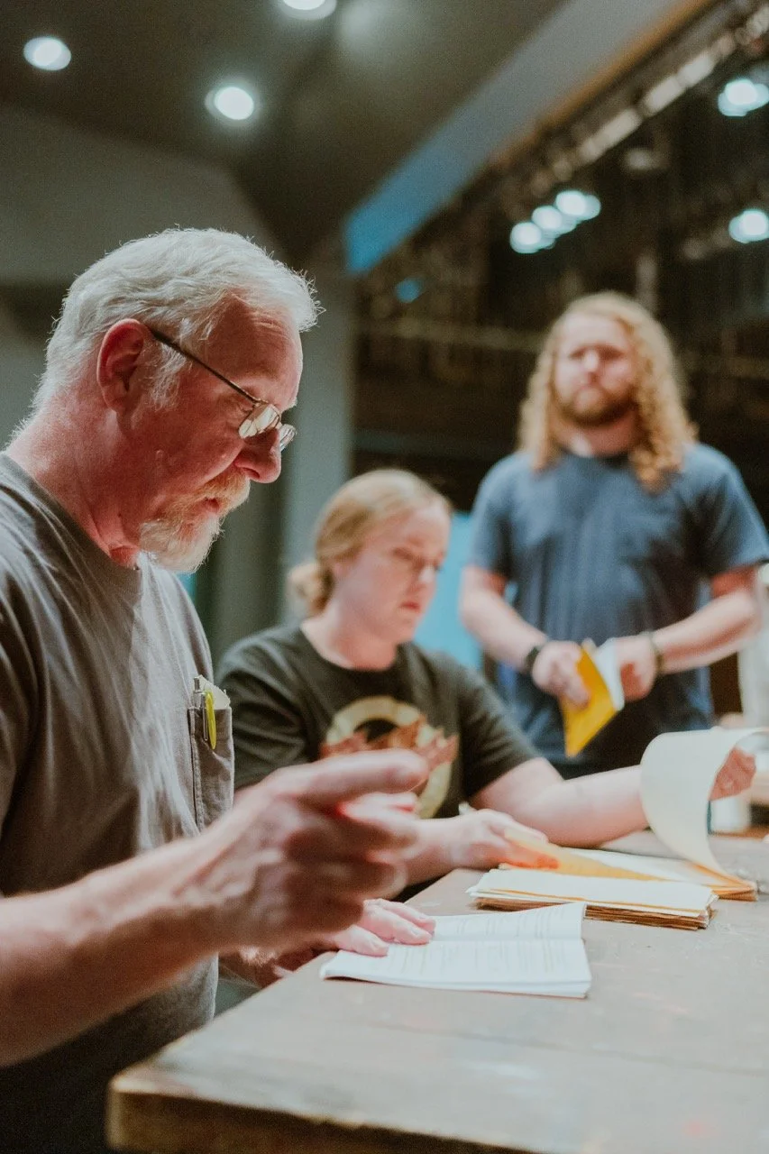 Three people engaged in a discussion or rehearsal, seated at a table with papers and a script in hand; the setting appears to be a theater or stage environment.
