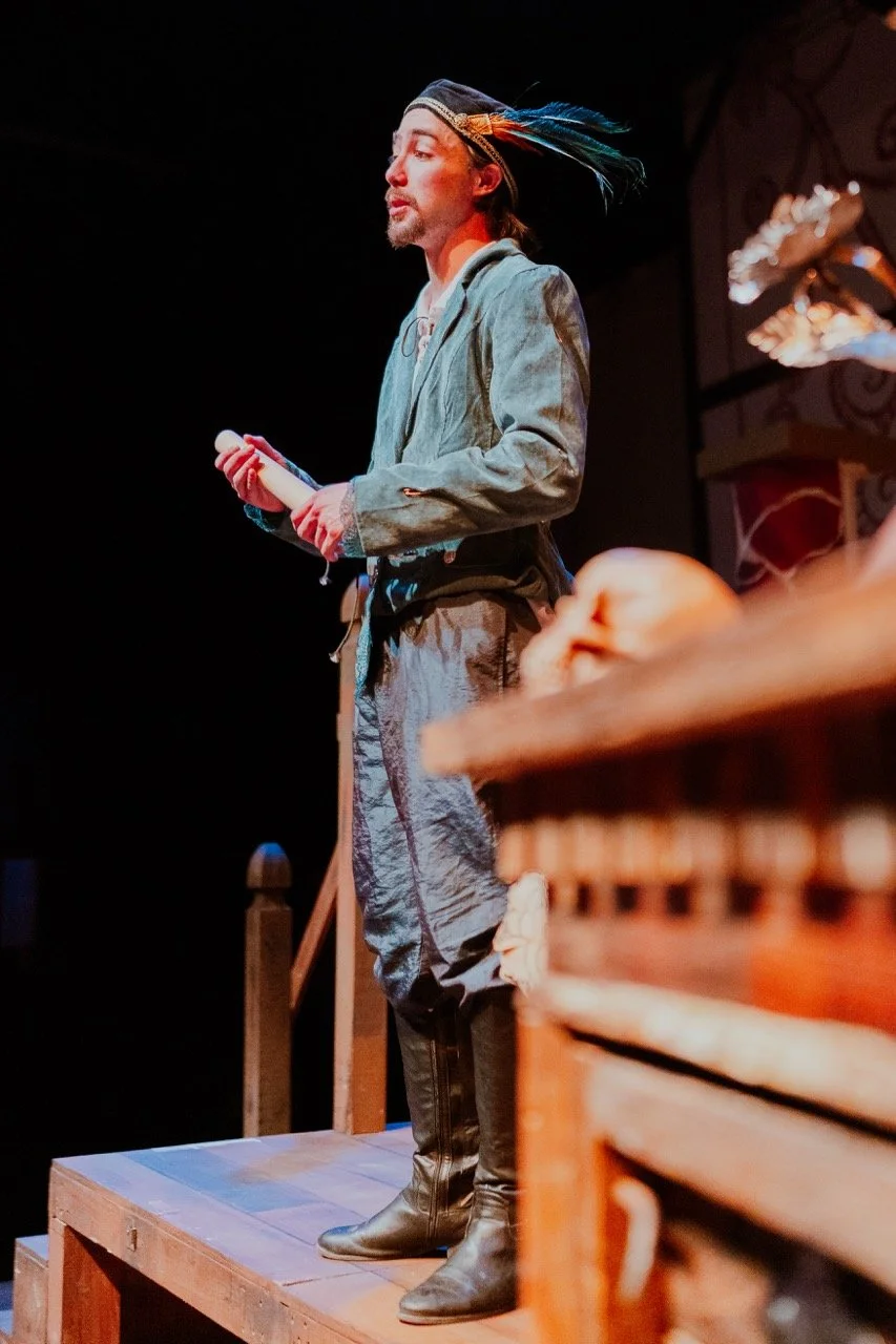 A person in historical costume, possibly a male actor, performing on stage with a rolled script in hand, wearing a hat with feathers.